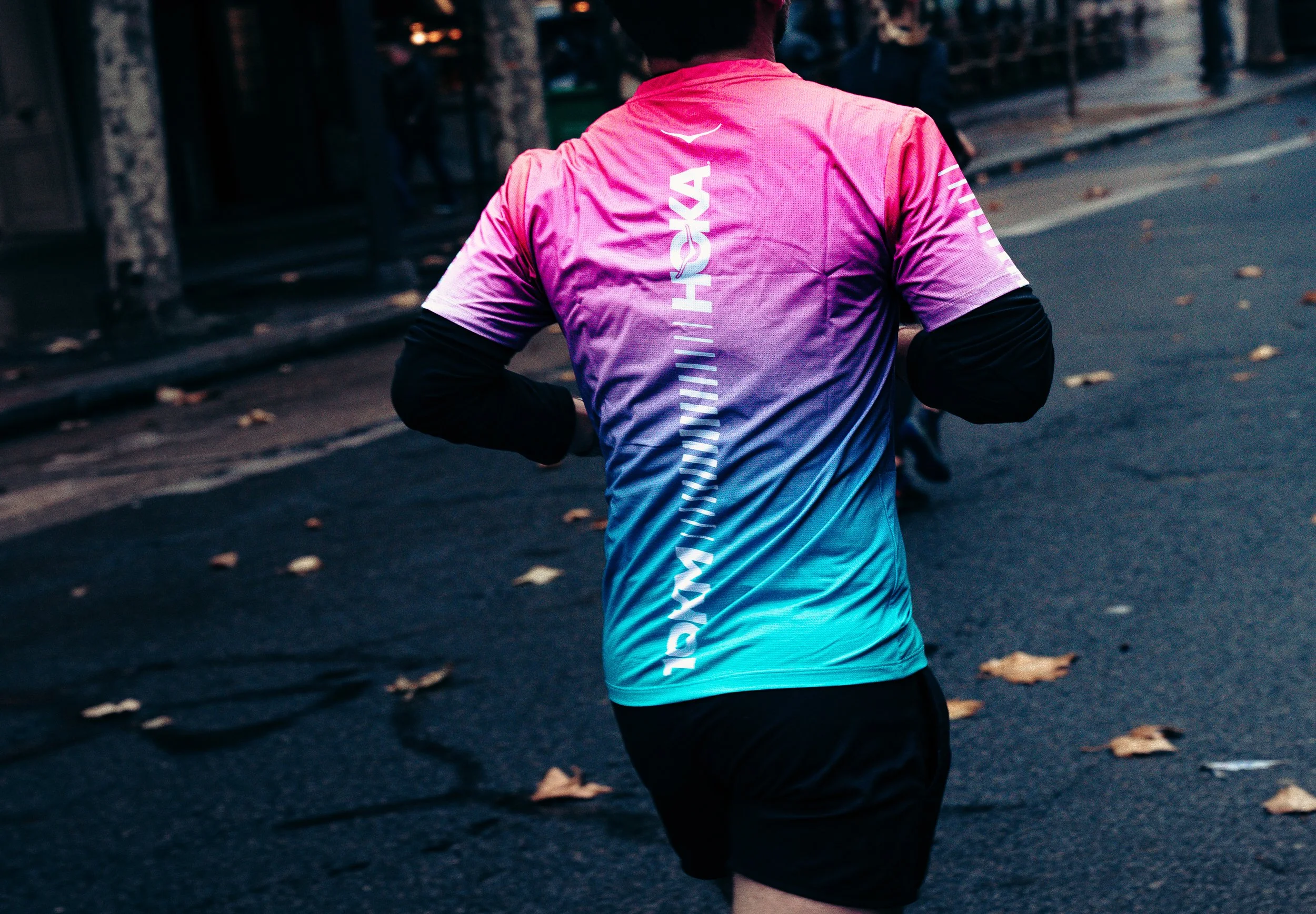 A person wearing a colorful Nike Marathon shirt running on a street with fallen leaves and a barrier on the sidewalk.