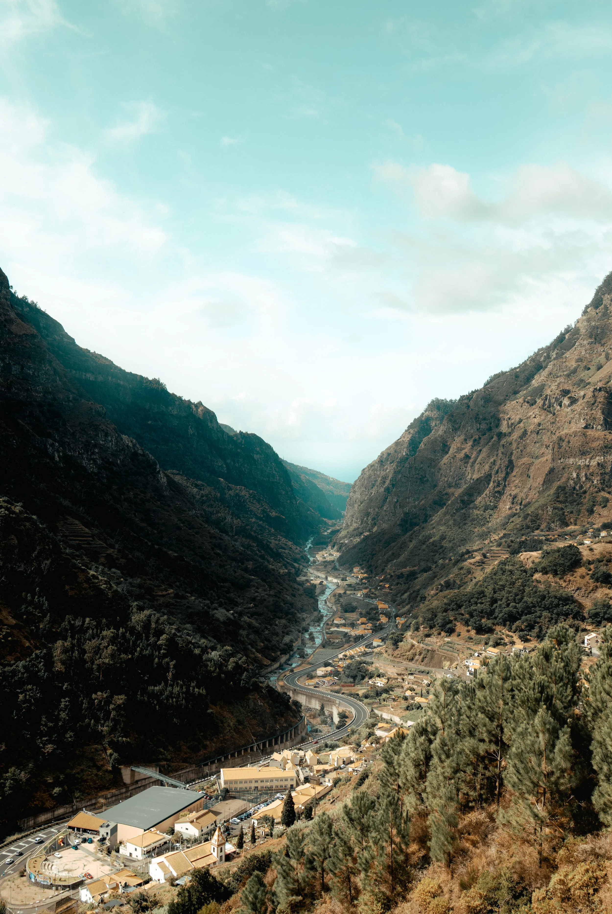 A scenic view of a winding road through a valley flanked by tall, steep mountains, with a small settlement and trees at the base.