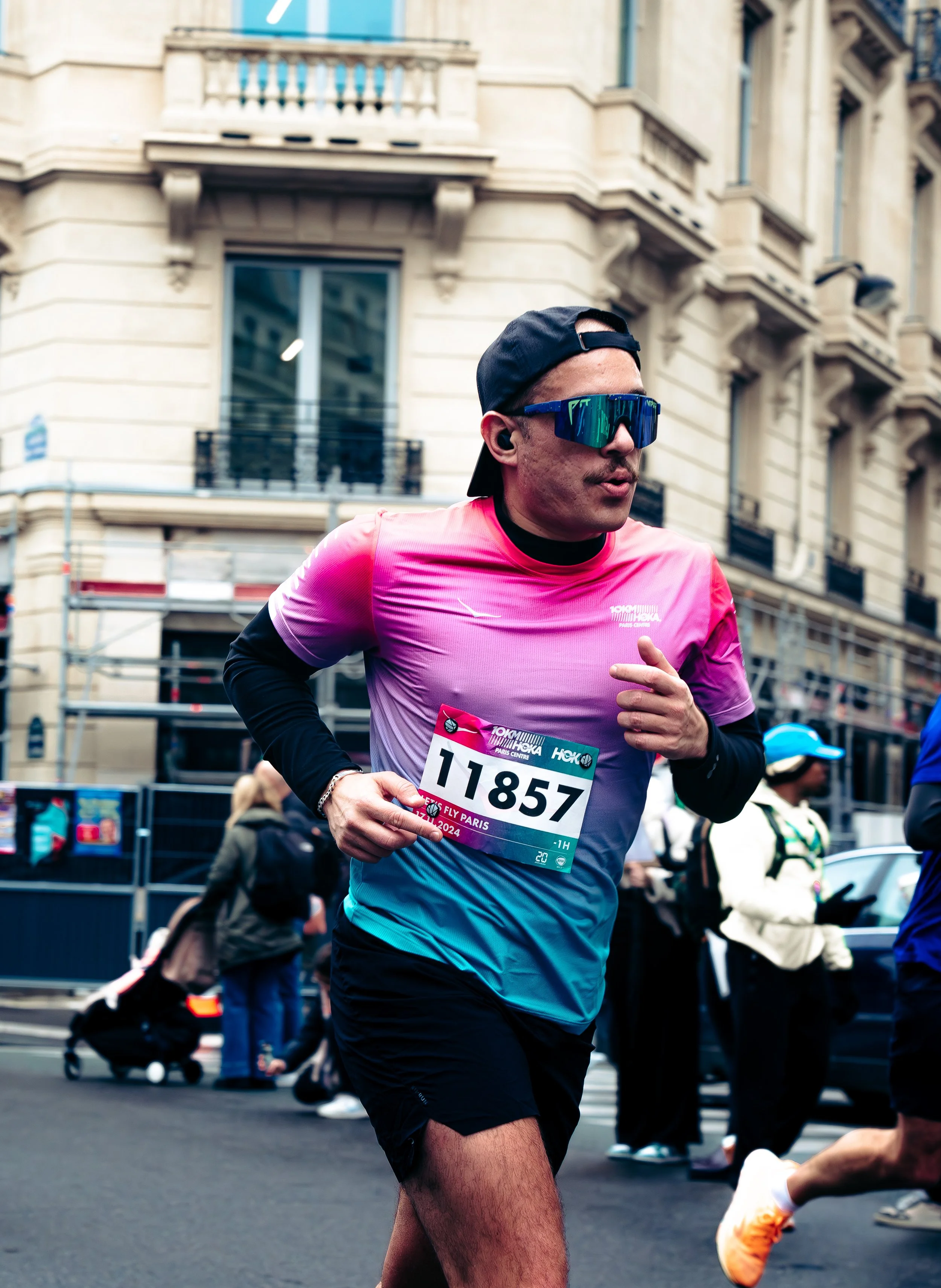 A male runner wearing sunglasses and a black cap participates in a marathon on a city street, with buildings and other runners in the background.