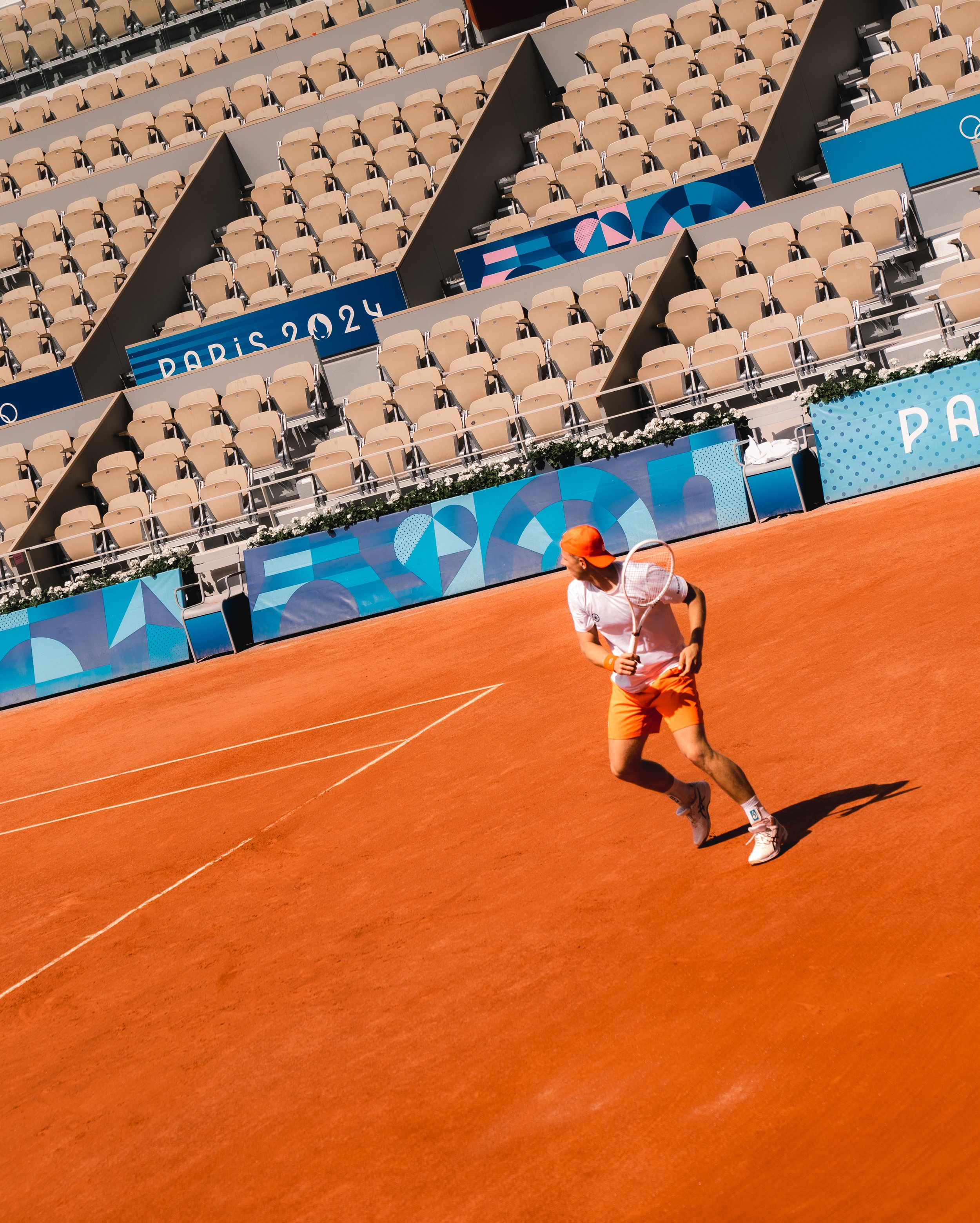 A tennis player in a white shirt, orange shorts, and white sneakers on a red clay court during the Paris 2024 Olympics, with empty spectator seats in the background.