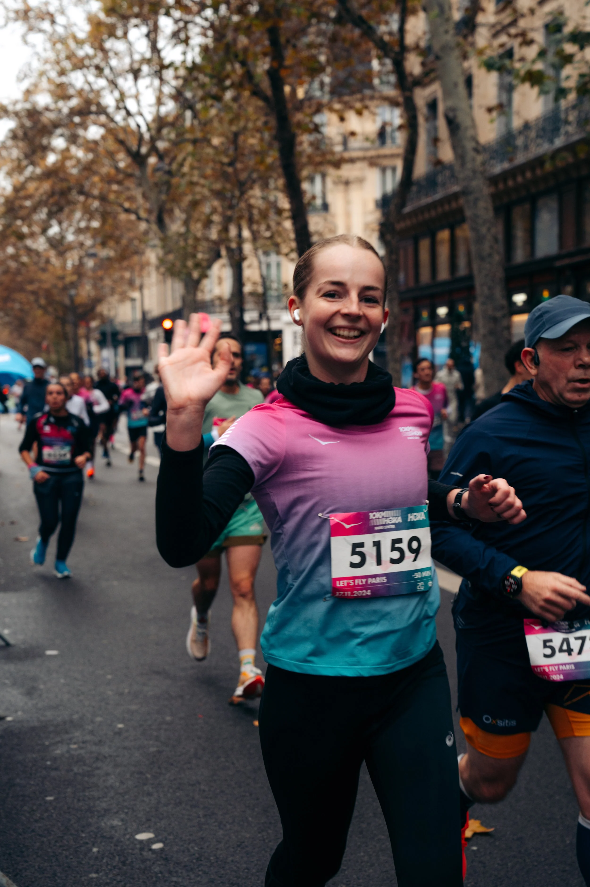 Smiling female marathon runner waving during race on city street lined with autumn trees and buildings, wearing a pink and blue running shirt with bib number 5159.