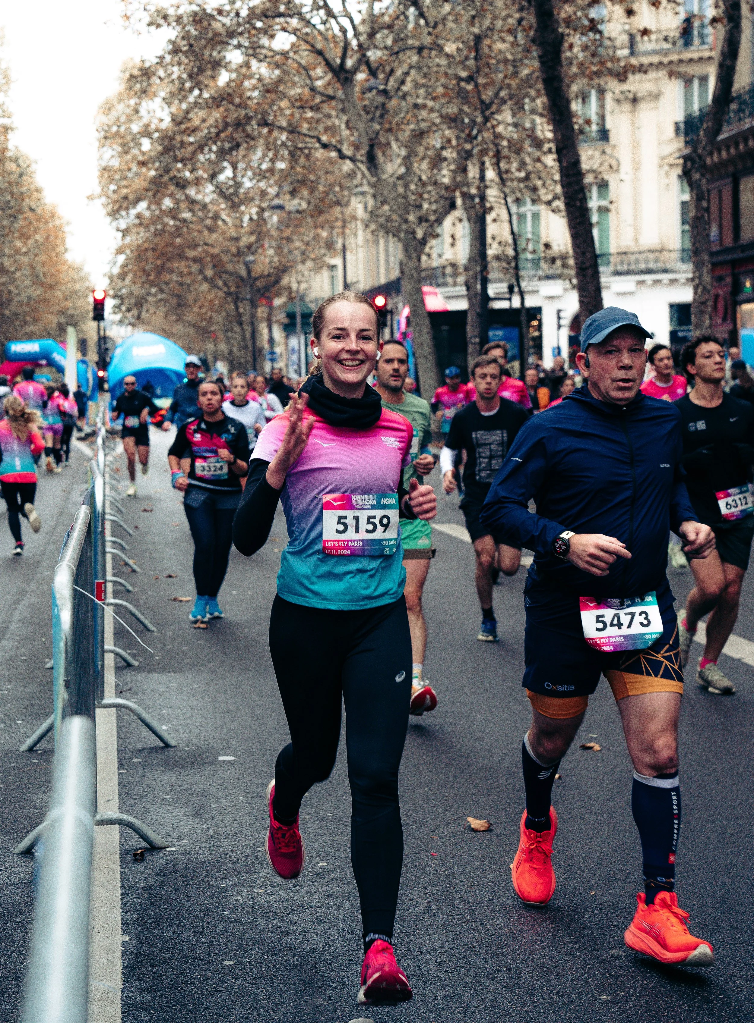 People participating in a marathon race on a city street, with runners wearing colorful athletic gear and race bibs, including a smiling woman in the foreground waving.