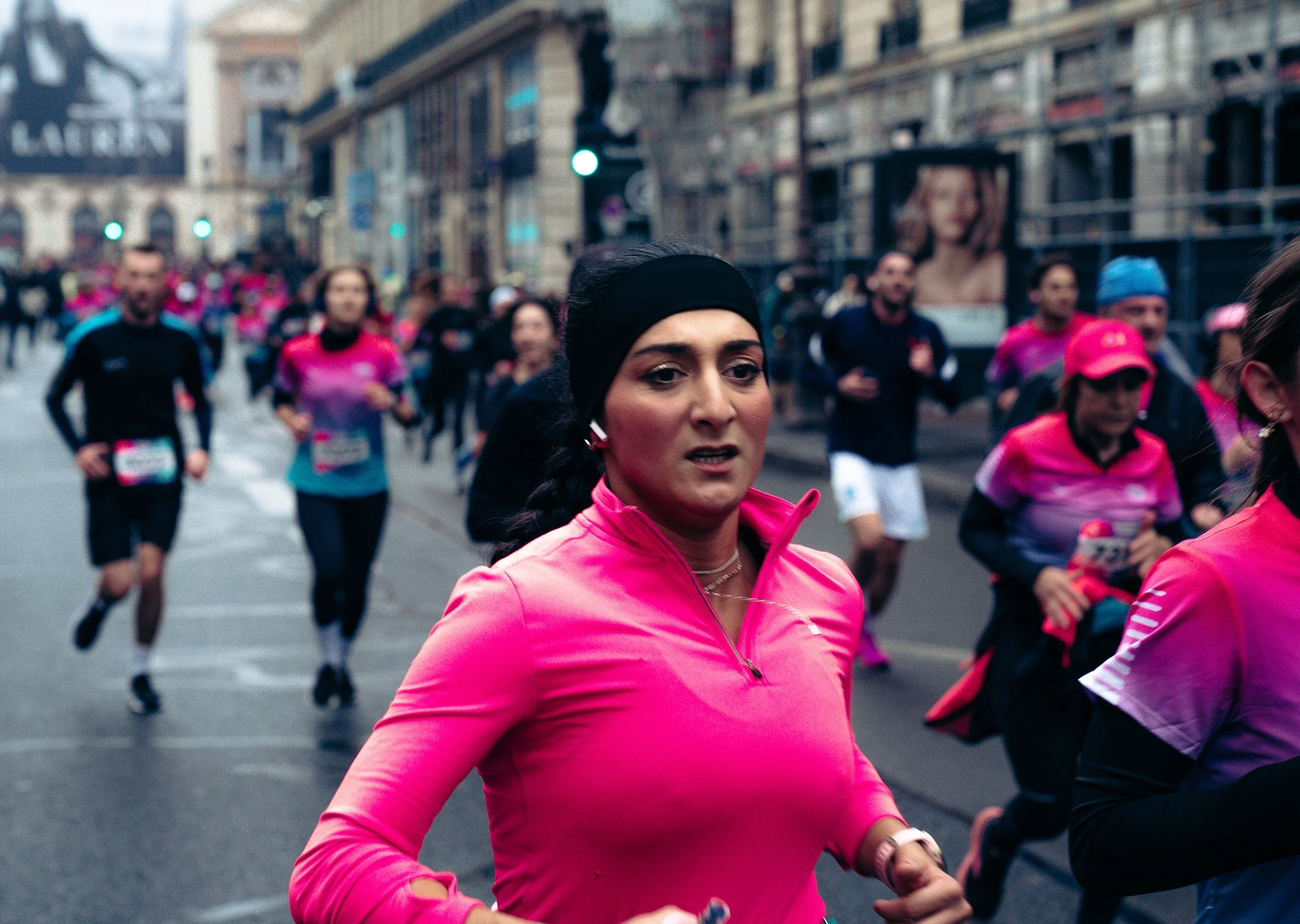 A group of runners participating in a marathon on a city street, with the woman in the foreground wearing a pink long-sleeve top and black headband.