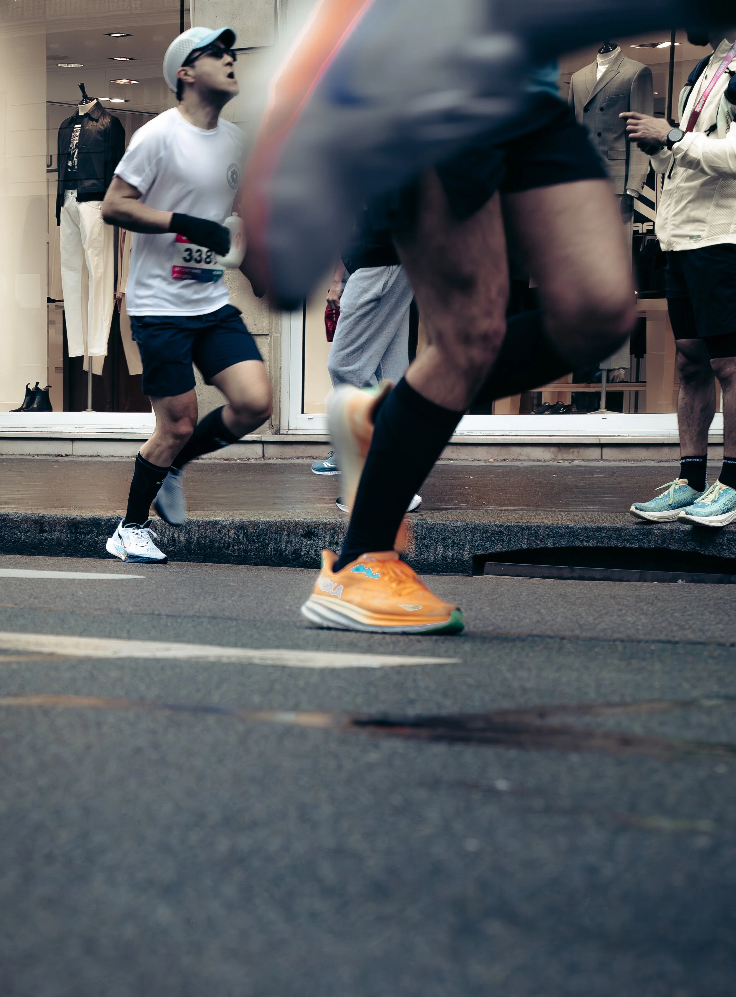 A man in a gray shirt and black shorts running down a city street during a race, with spectators and storefronts in the background.