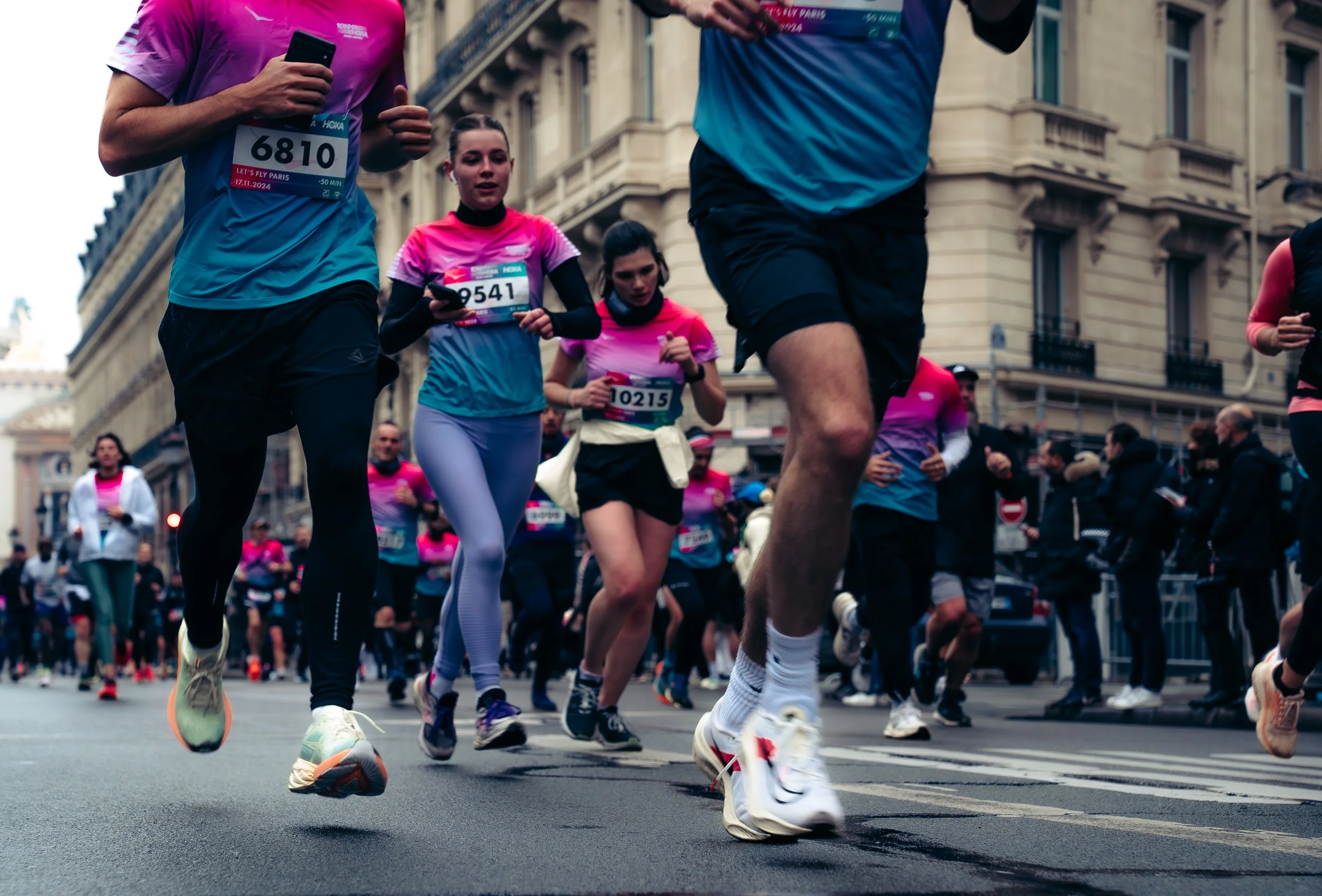 People participating in a road marathon, running on city street with historic buildings, wearing colorful athletic clothing and race bibs.