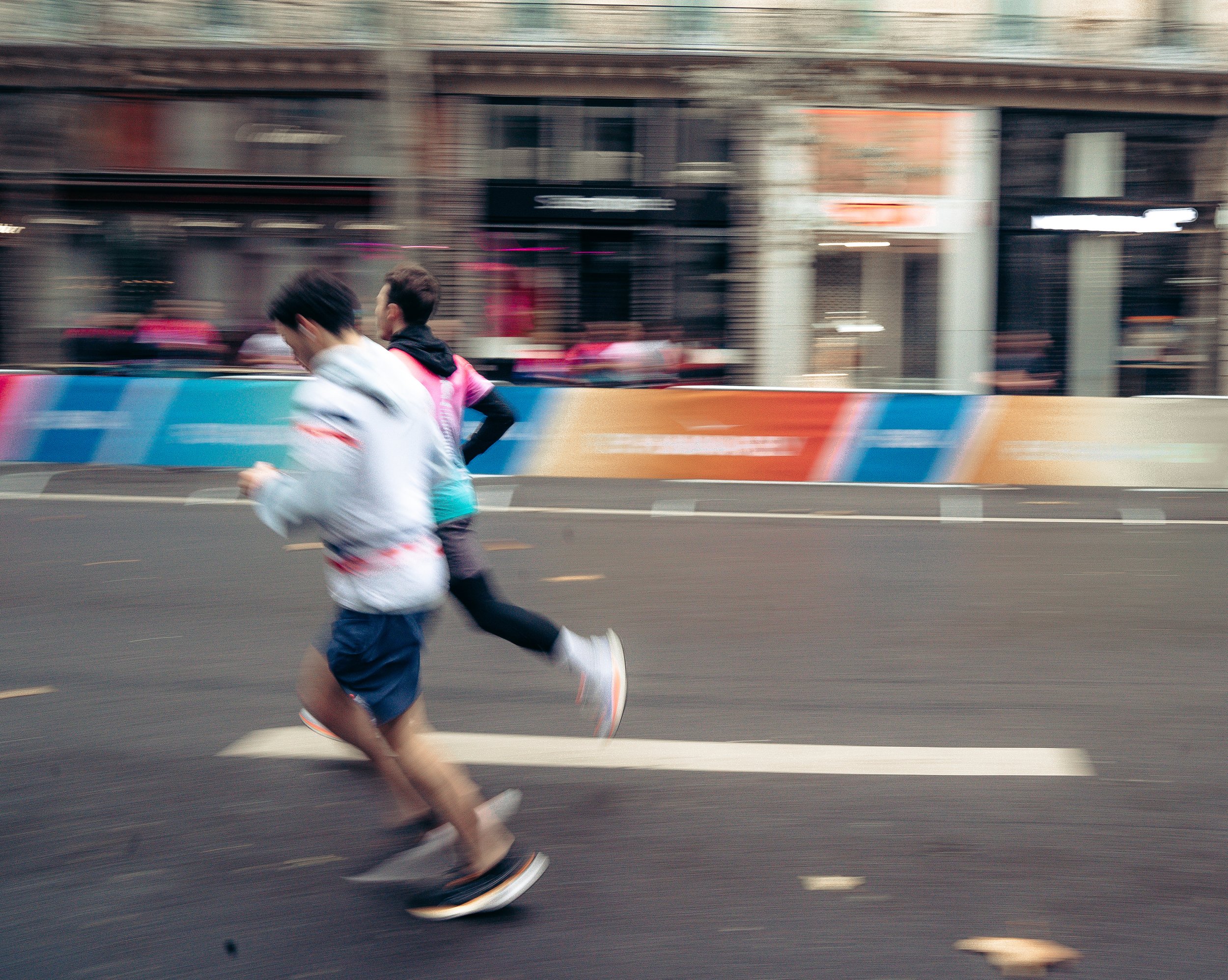 Two runners running on a city street with blurred buildings and barriers in the background.