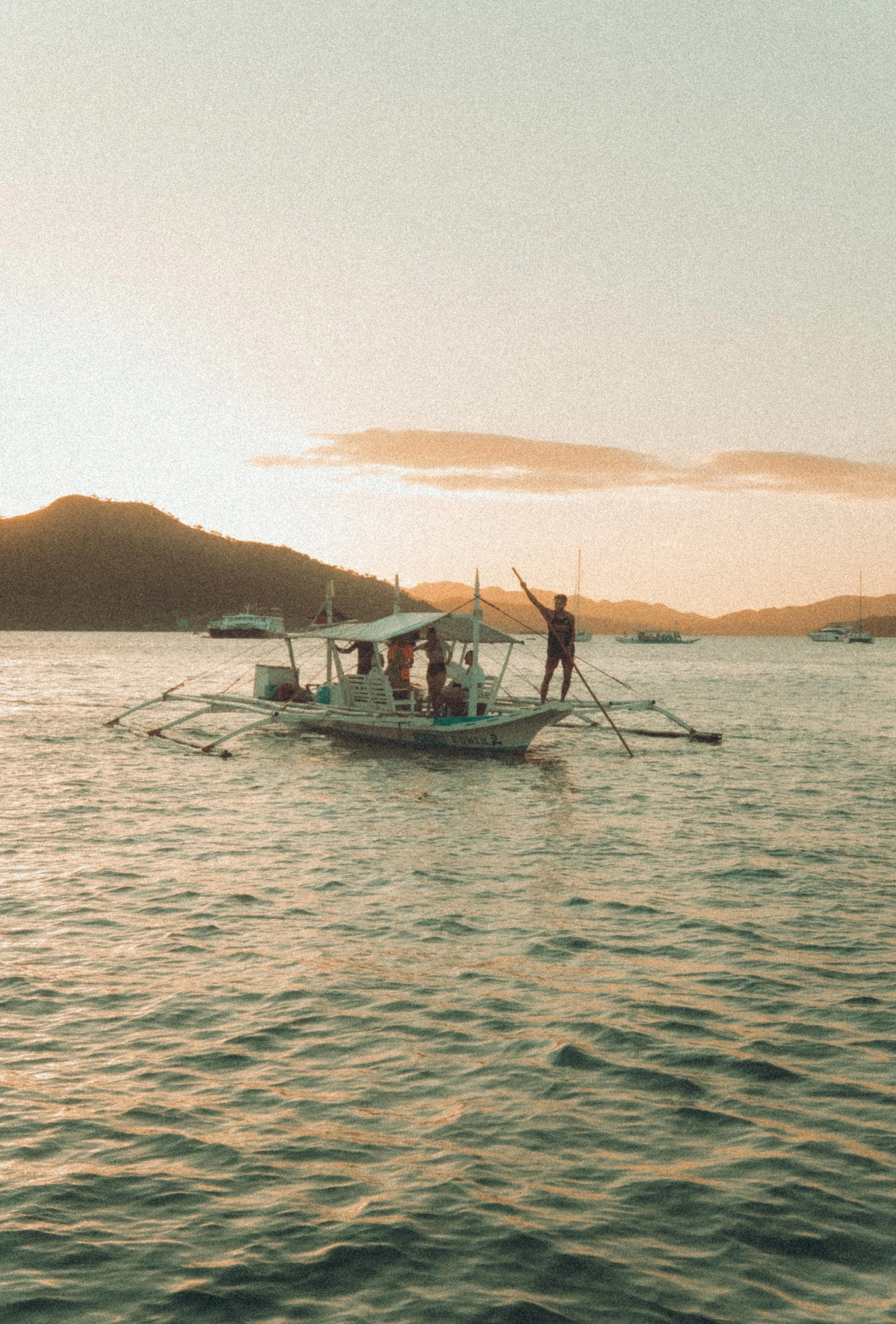 A boat with people on it, with one person standing and using a pole, in a calm body of water during sunset, with mountains in the background.