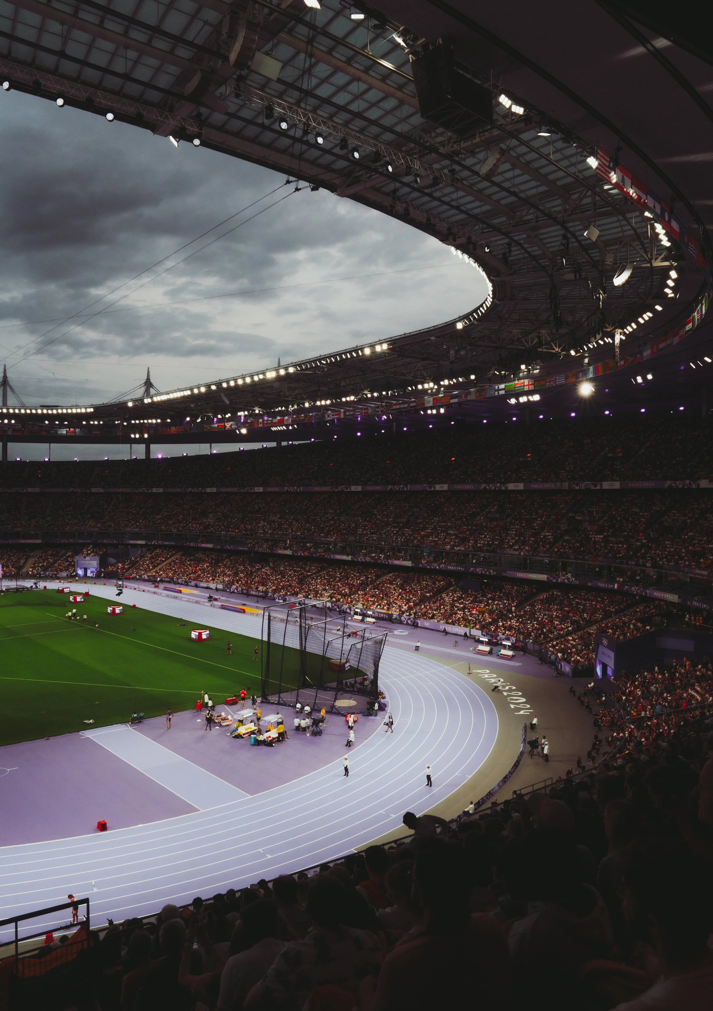 Inside a large stadium with a track and field event setup, an empty running track surrounds a grassy field, with many spectators in the stands and cloudy skies overhead.