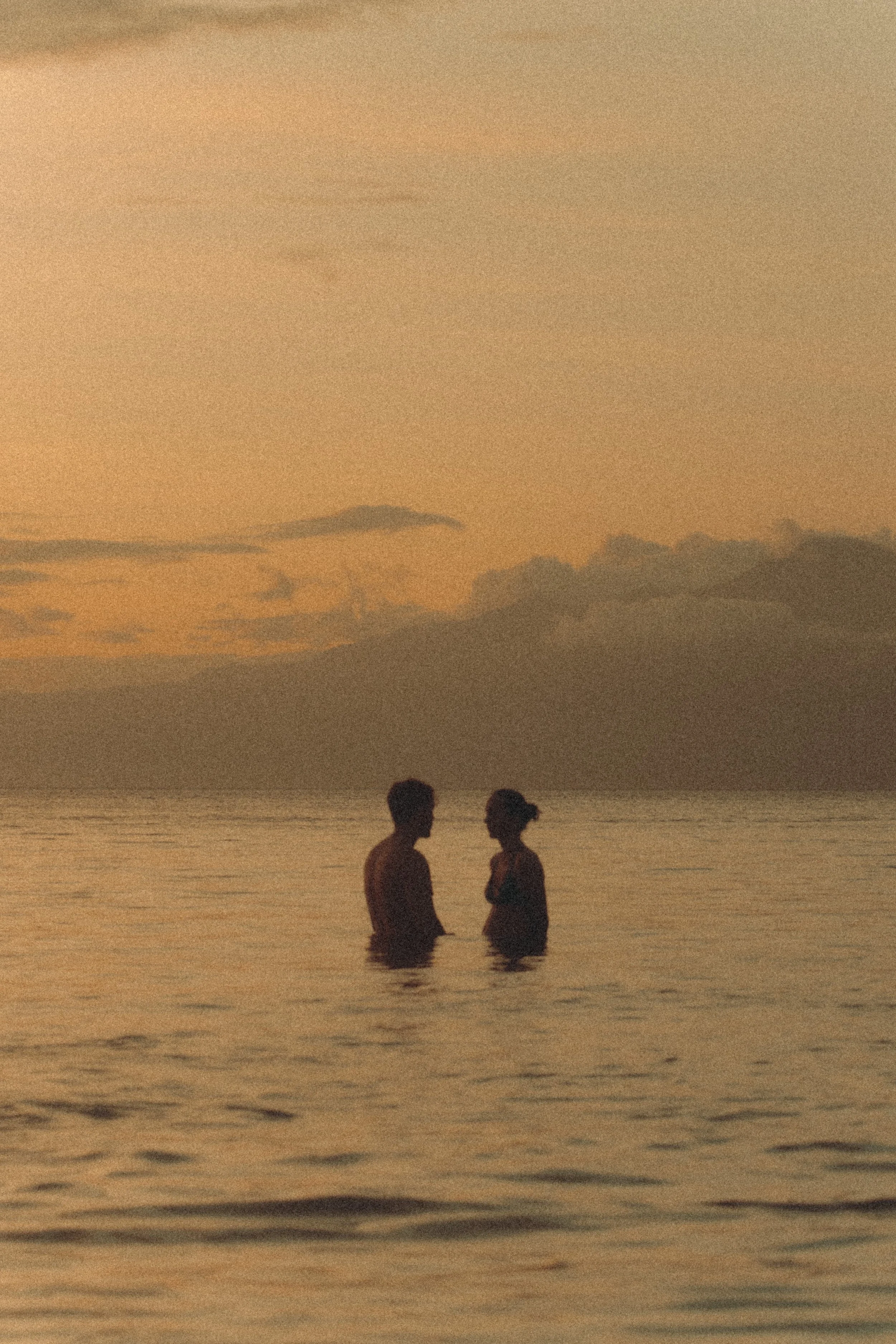 A couple standing in the ocean at sunset, facing each other, with a distant mountain and cloudy sky in the background.