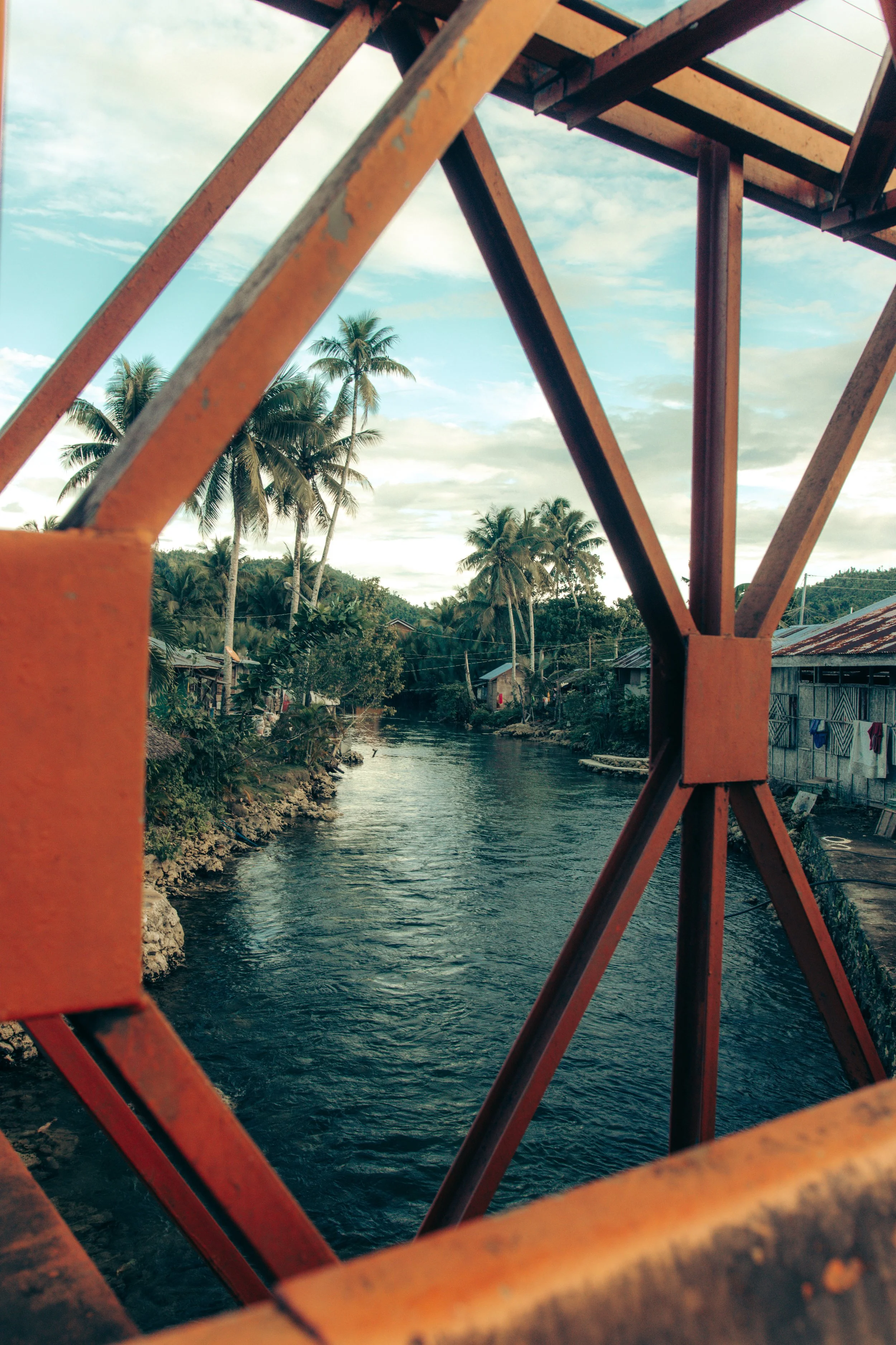View of a river with houses and palm trees on either side, taken from a bridge with rusted metal railings.