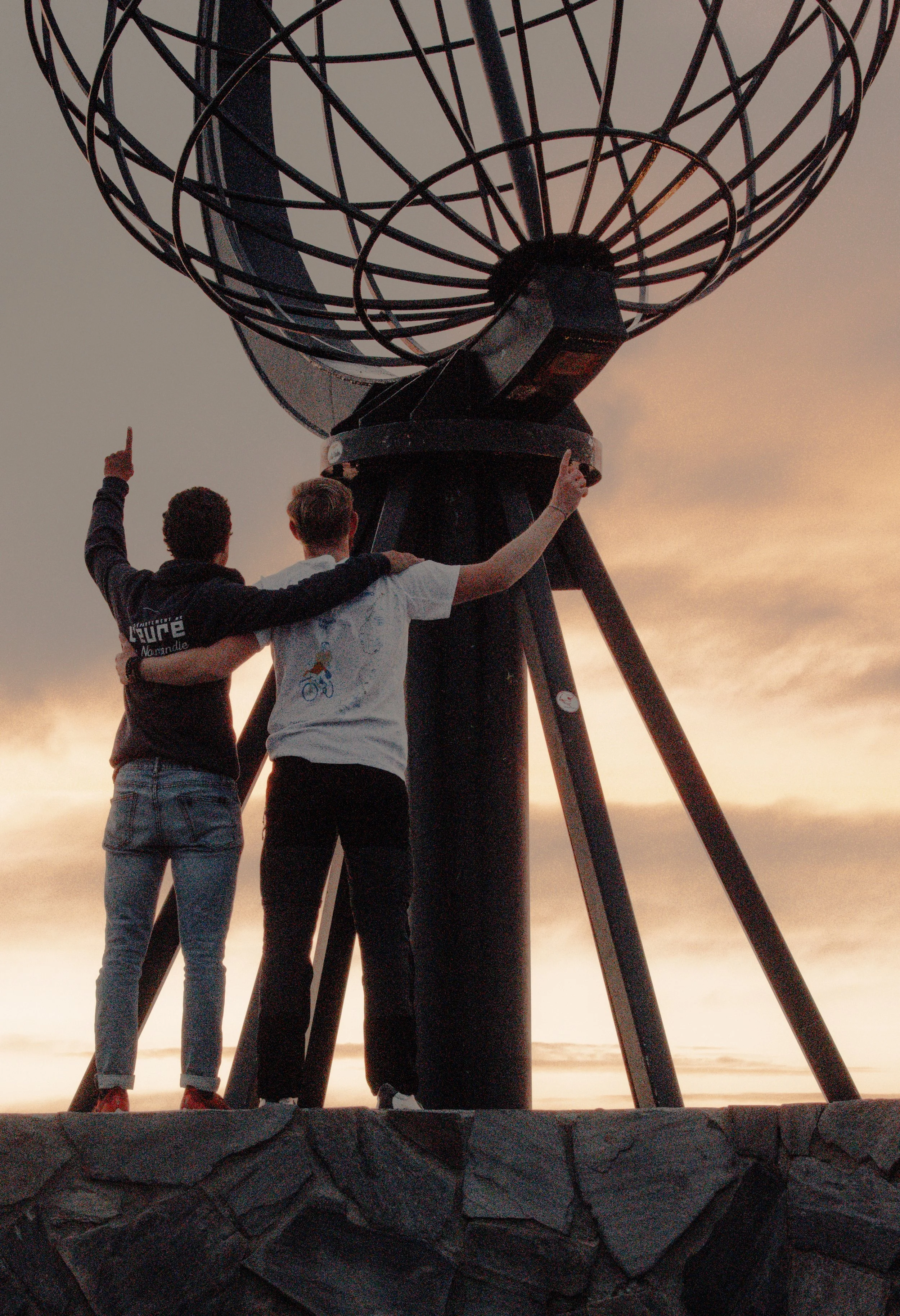 Two people standing with their arms around each other, pointing upwards, in front of a large globe monument at sunset.