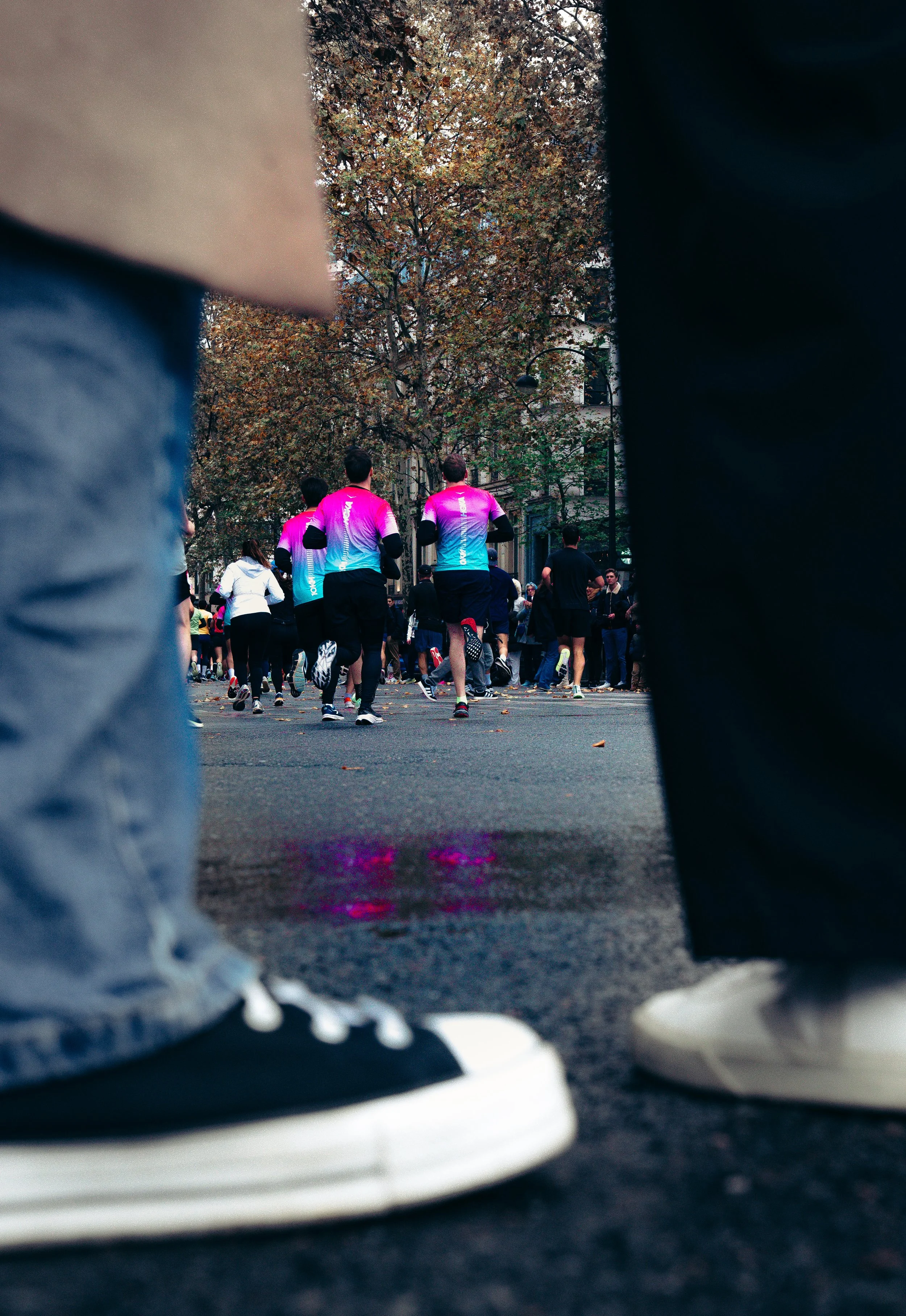 View of a group of people running in a race, photographed from the ground between two pairs of legs and feet, with a tree-lined street and autumn foliage in the background.