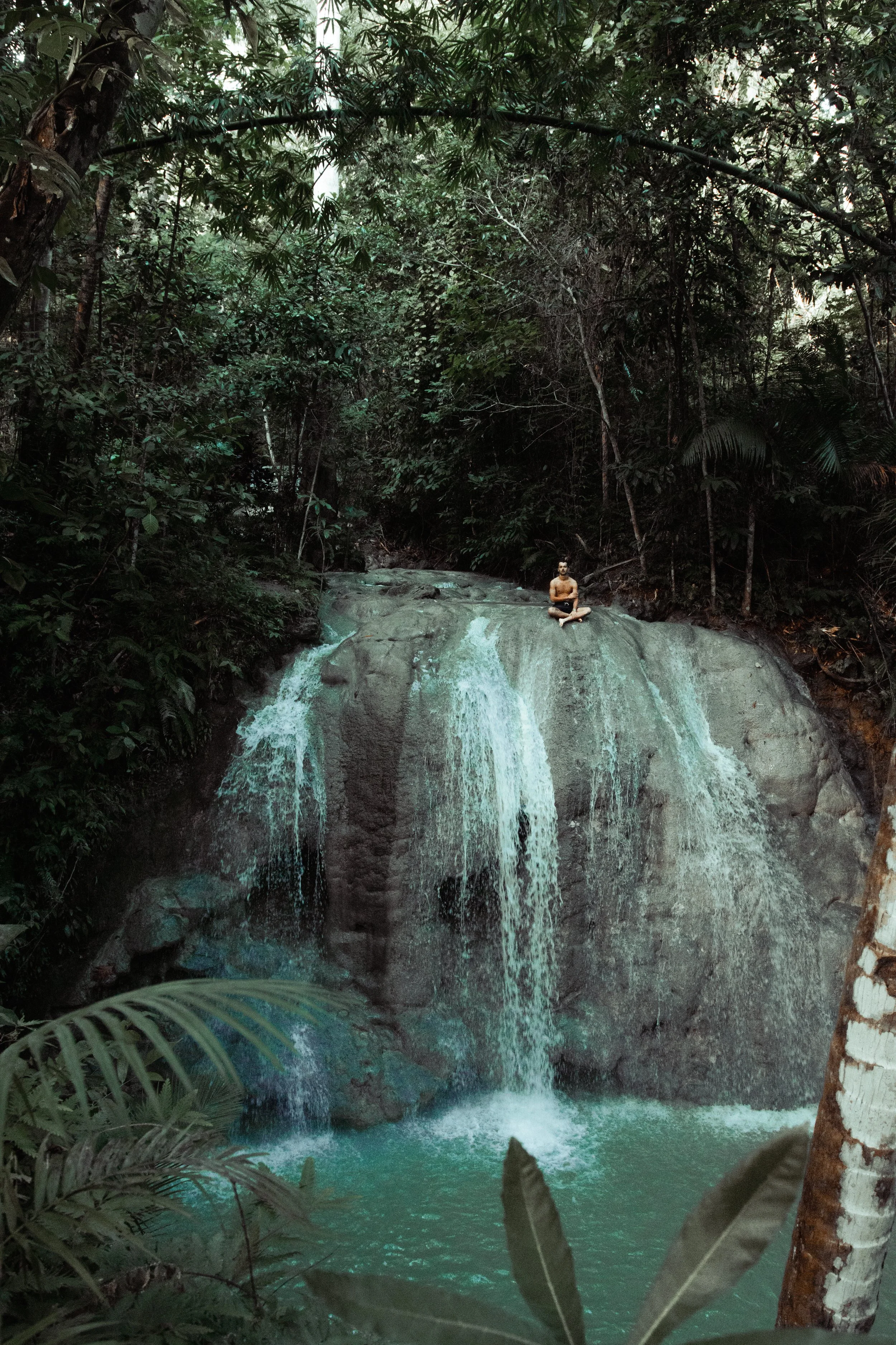 A person meditating on a large rock underneath a waterfall in a lush, green rainforest.