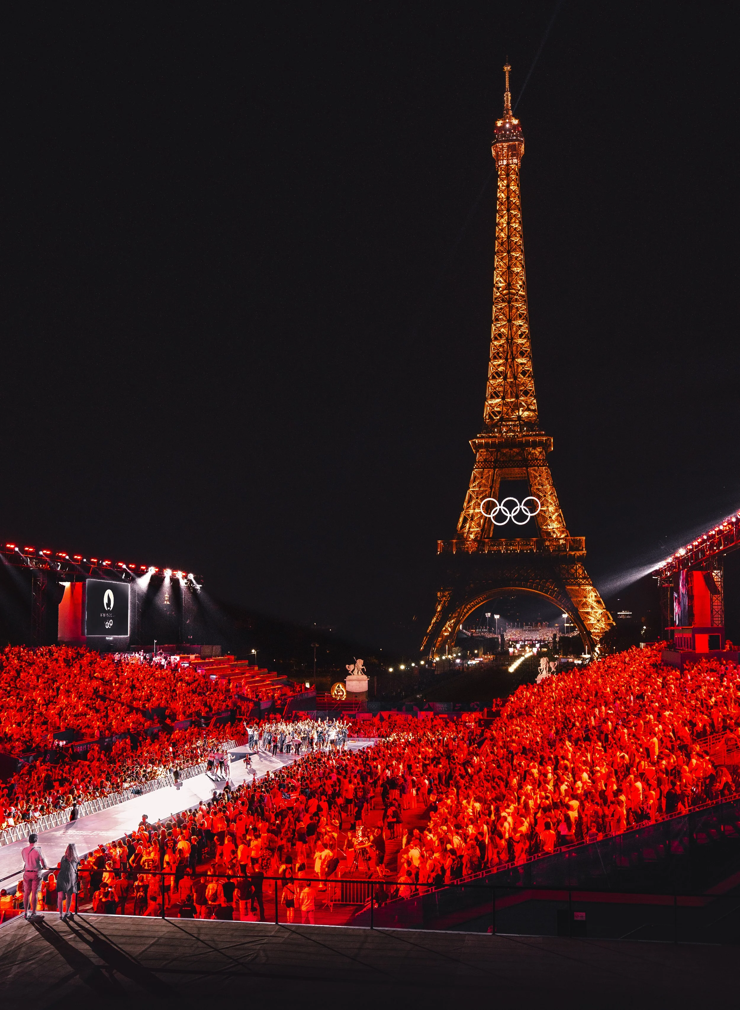 Nighttime scene of the Eiffel Tower illuminated with the Olympic rings, with a large crowd dressed in red gathered in front for an event. The Eiffel Tower is lit with golden lights, and the Olympic rings are prominently displayed on its middle.