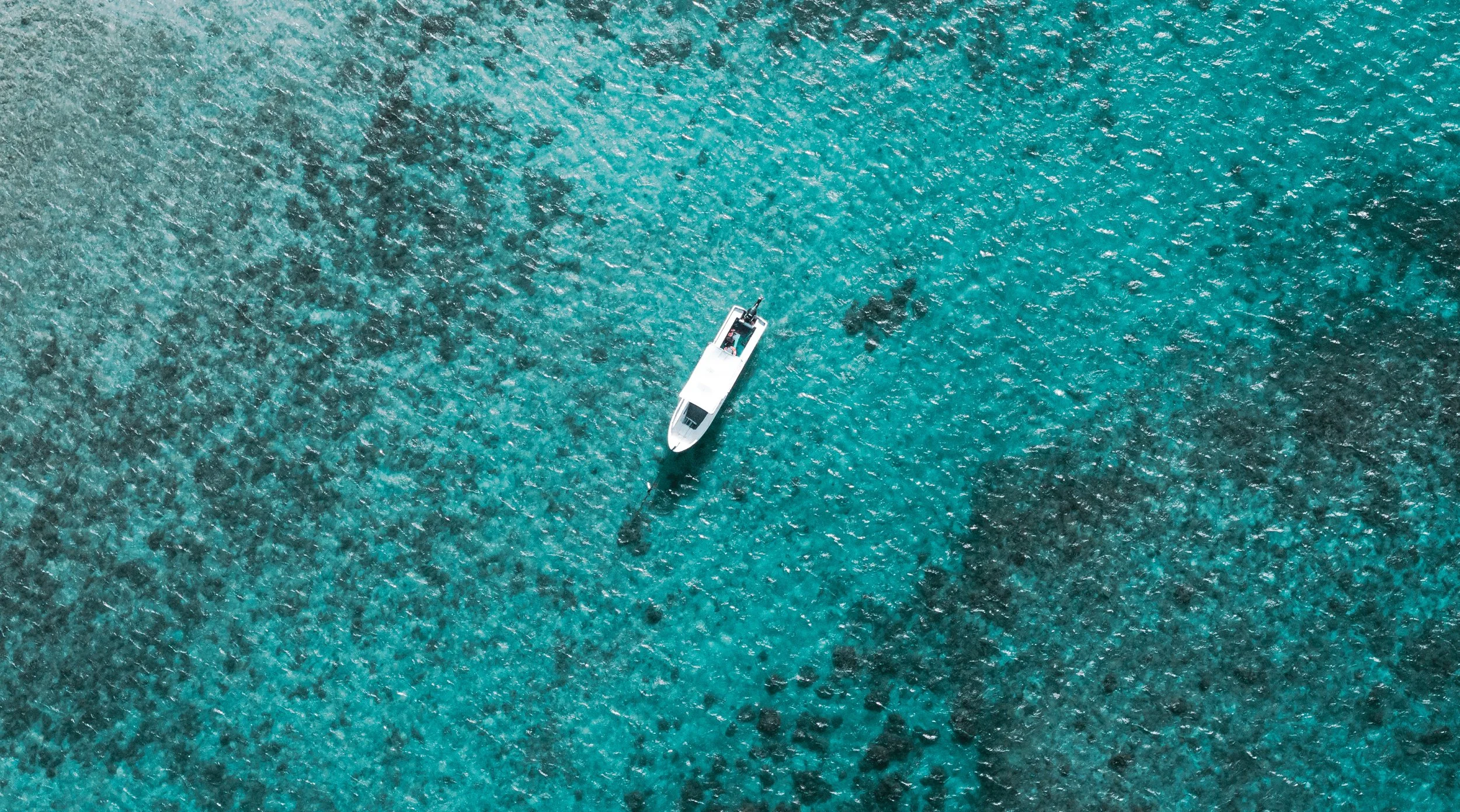 Aerial view of a white boat on clear blue ocean water with dark rocks below the surface.