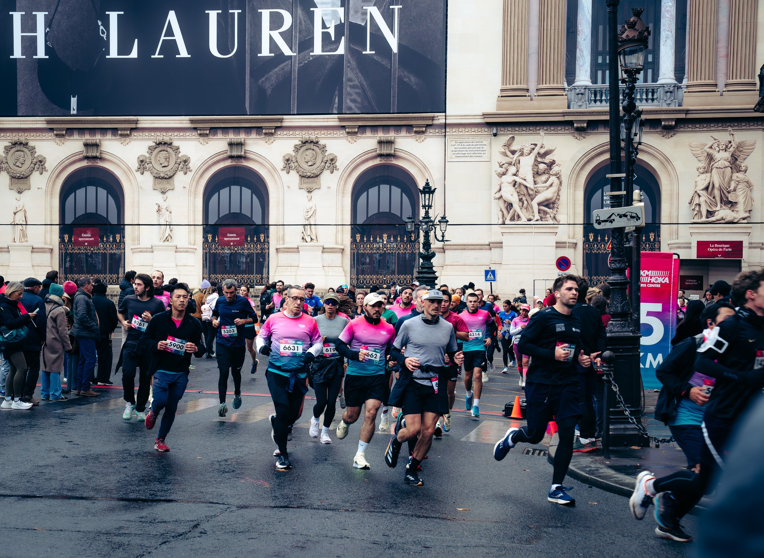 A group of runners participating in a race on a city street, passing by an ornate building with classical architectural features and sculptures, with spectators on the sidewalk.