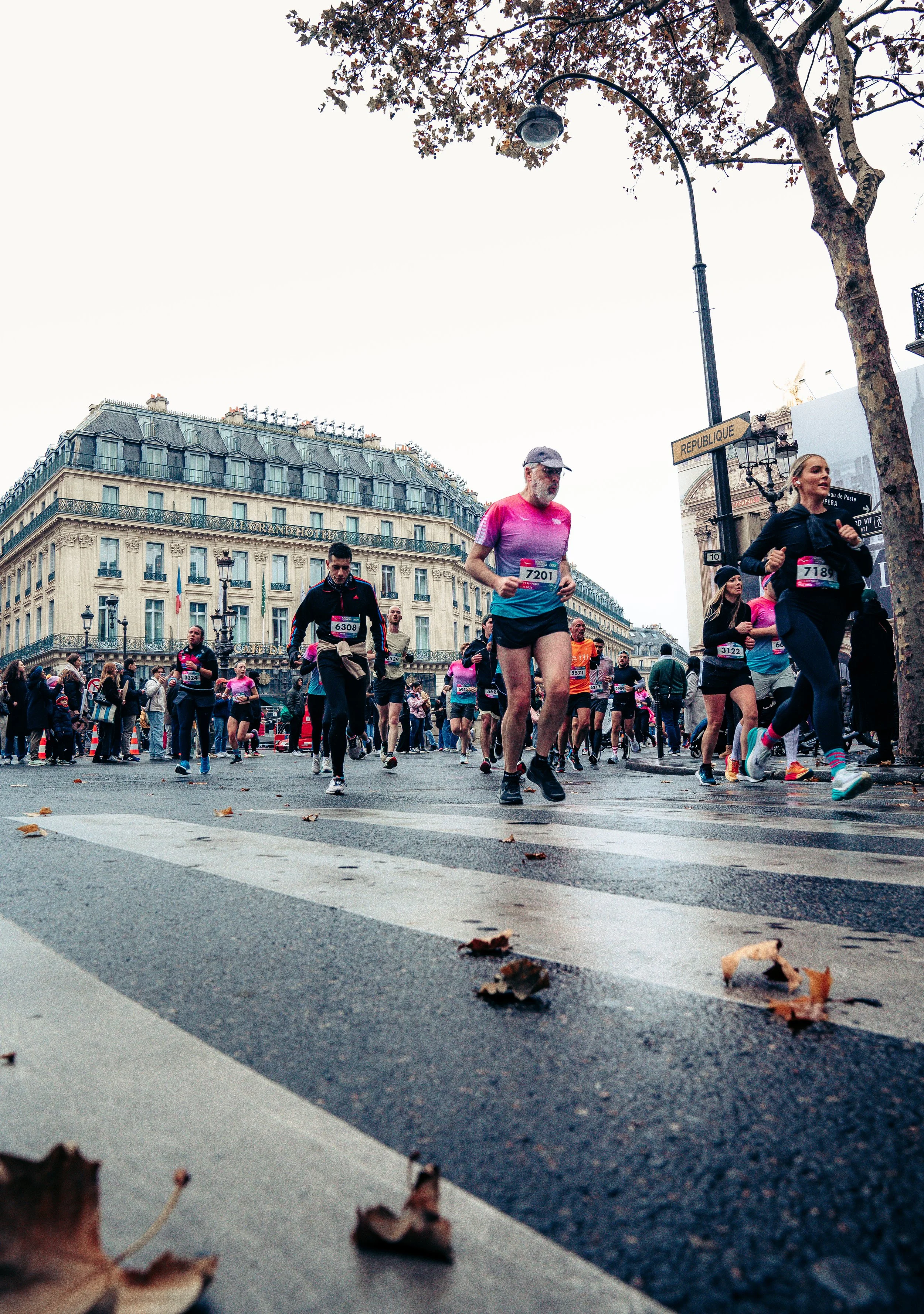 People participating in a marathon race running through a city street with historic buildings in the background and fallen leaves on the pavement.