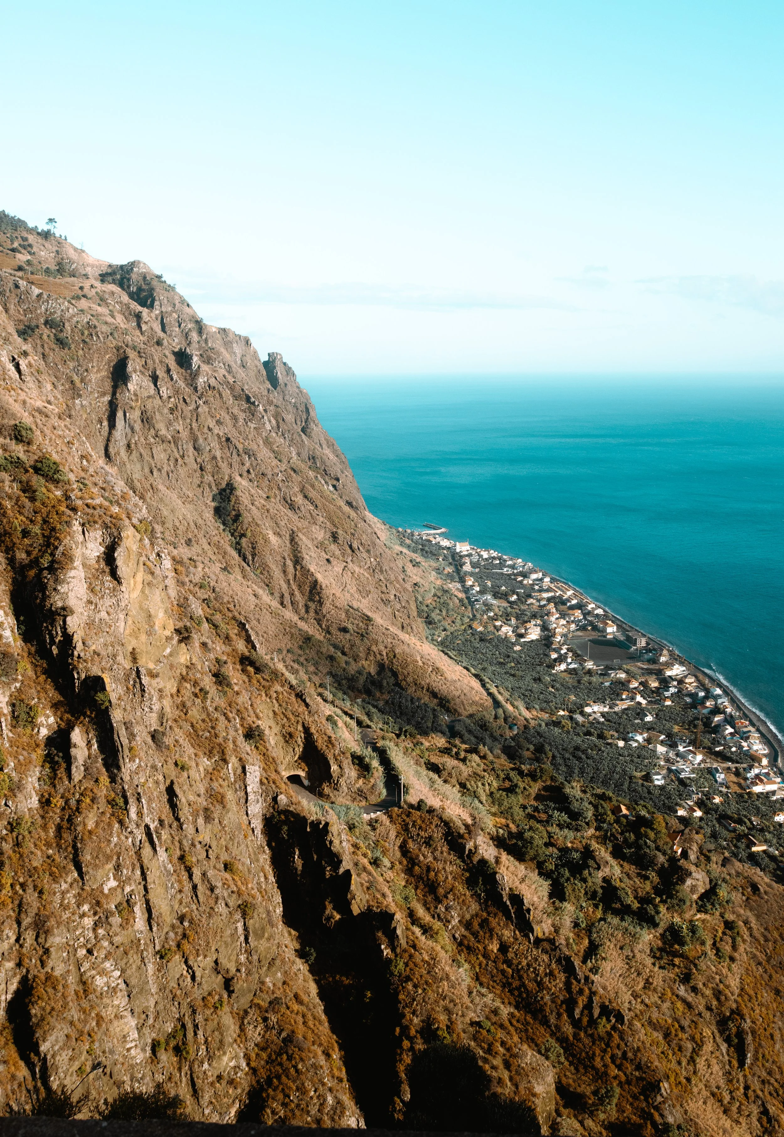A coastal landscape with steep mountains, a town along the shoreline, and the ocean extending into the horizon under a light blue sky.