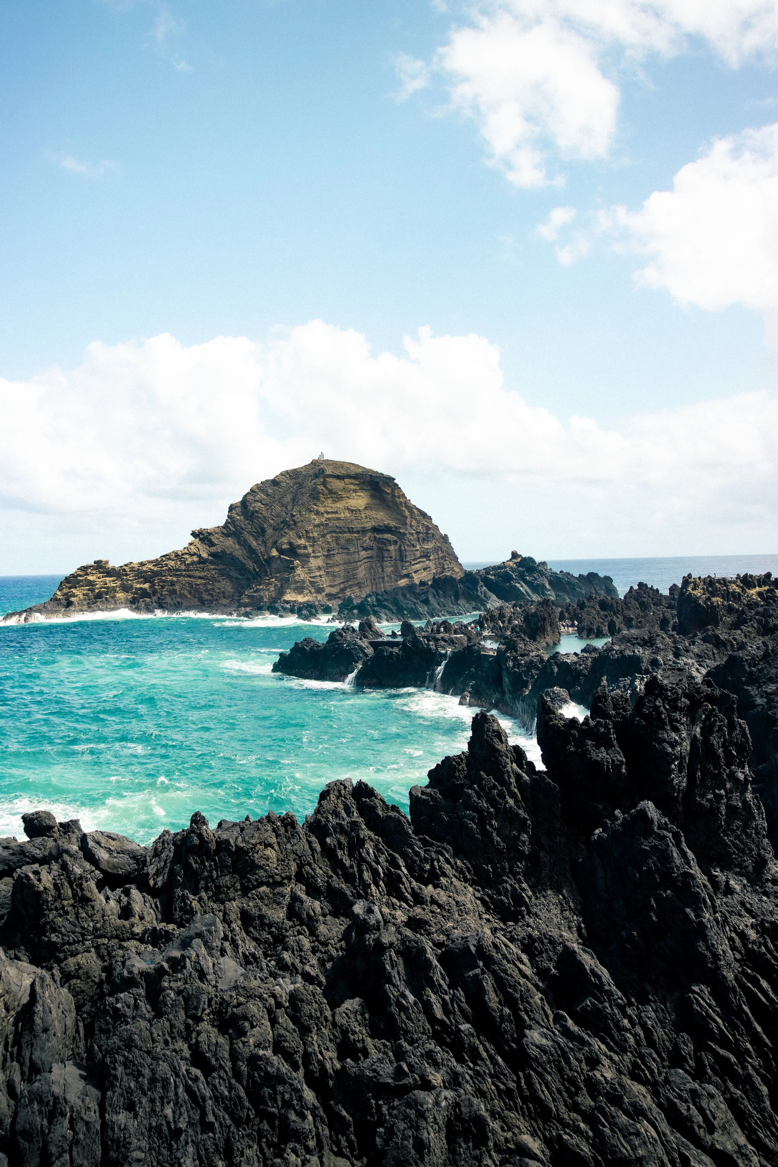 A rocky coastal landscape with black volcanic rocks in the foreground, turquoise waves crashing against the rocks, and a large island or rock formation in the distance under a partly cloudy sky.