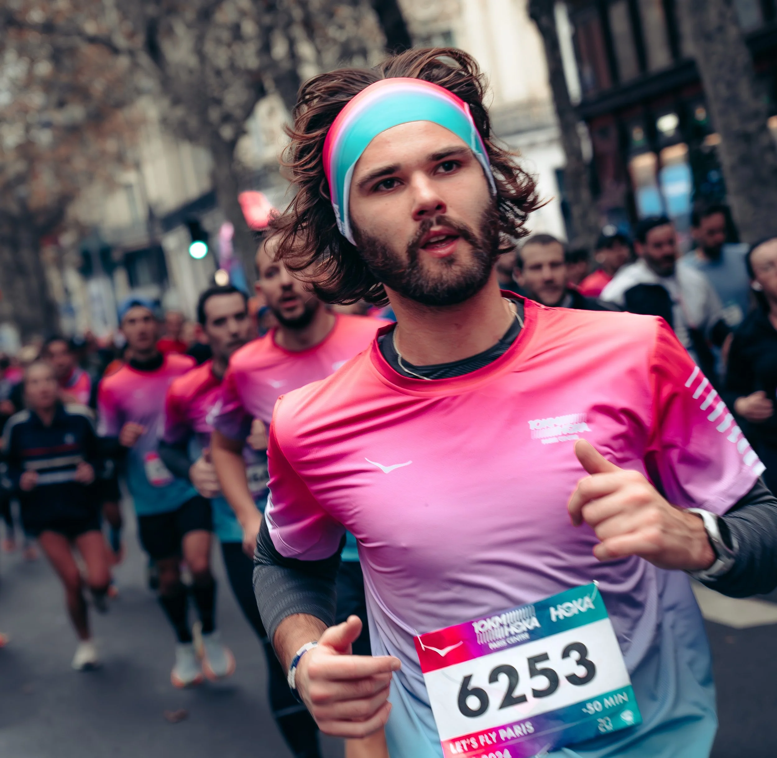 A man with long hair, beard, and colorful headband running in a marathon, wearing a pink and white athletic shirt and numbered bib, with other runners behind him on a city street.