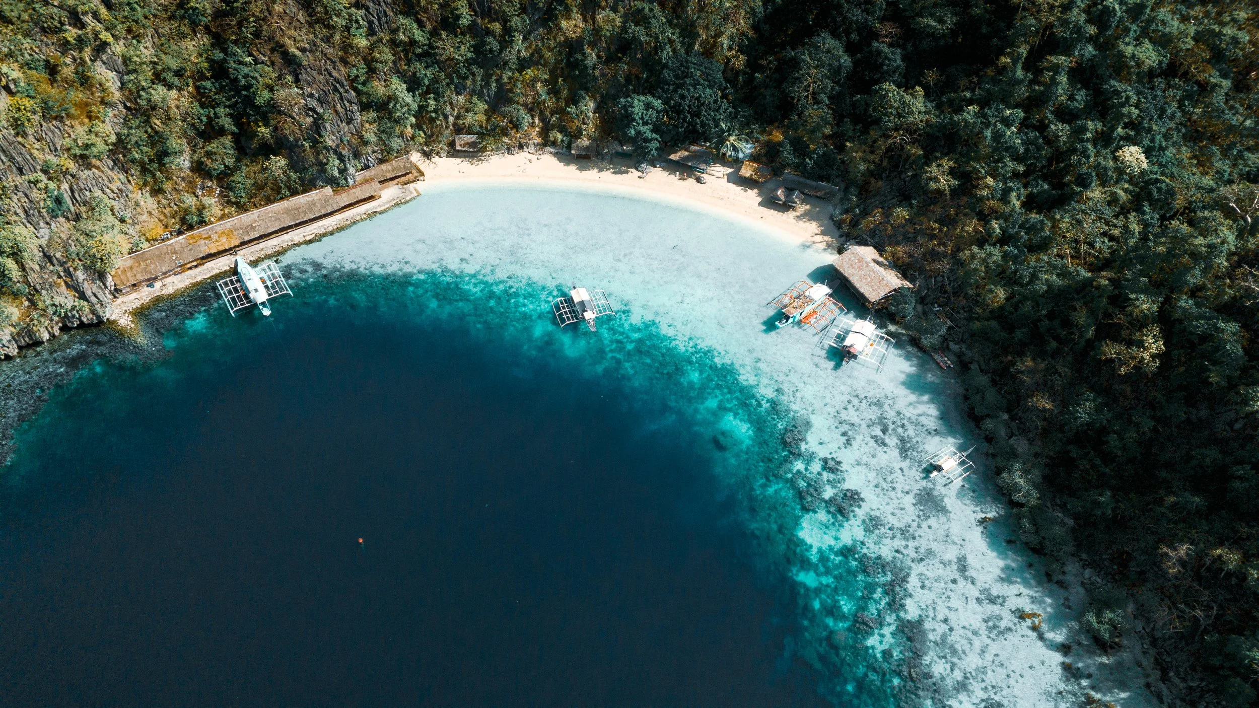 Aerial view of a tropical beach with white sand, surrounded by dense green forest, and calm turquoise waters, with several small boats docked near the shoreline.