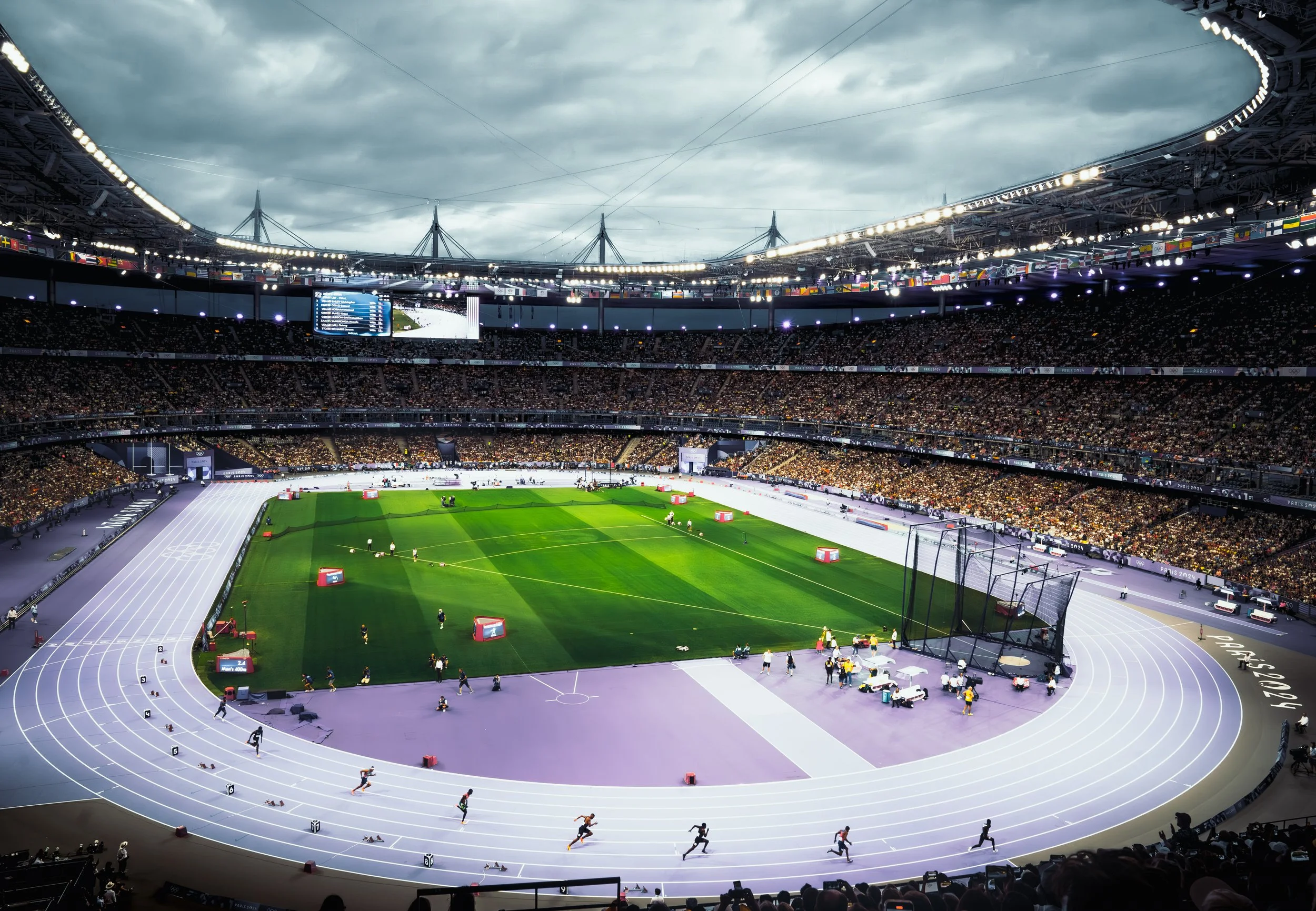 Olympic stadium with a running track and a green field, multiple athletes running on the track, large audience in the stands, overcast sky above the stadium.