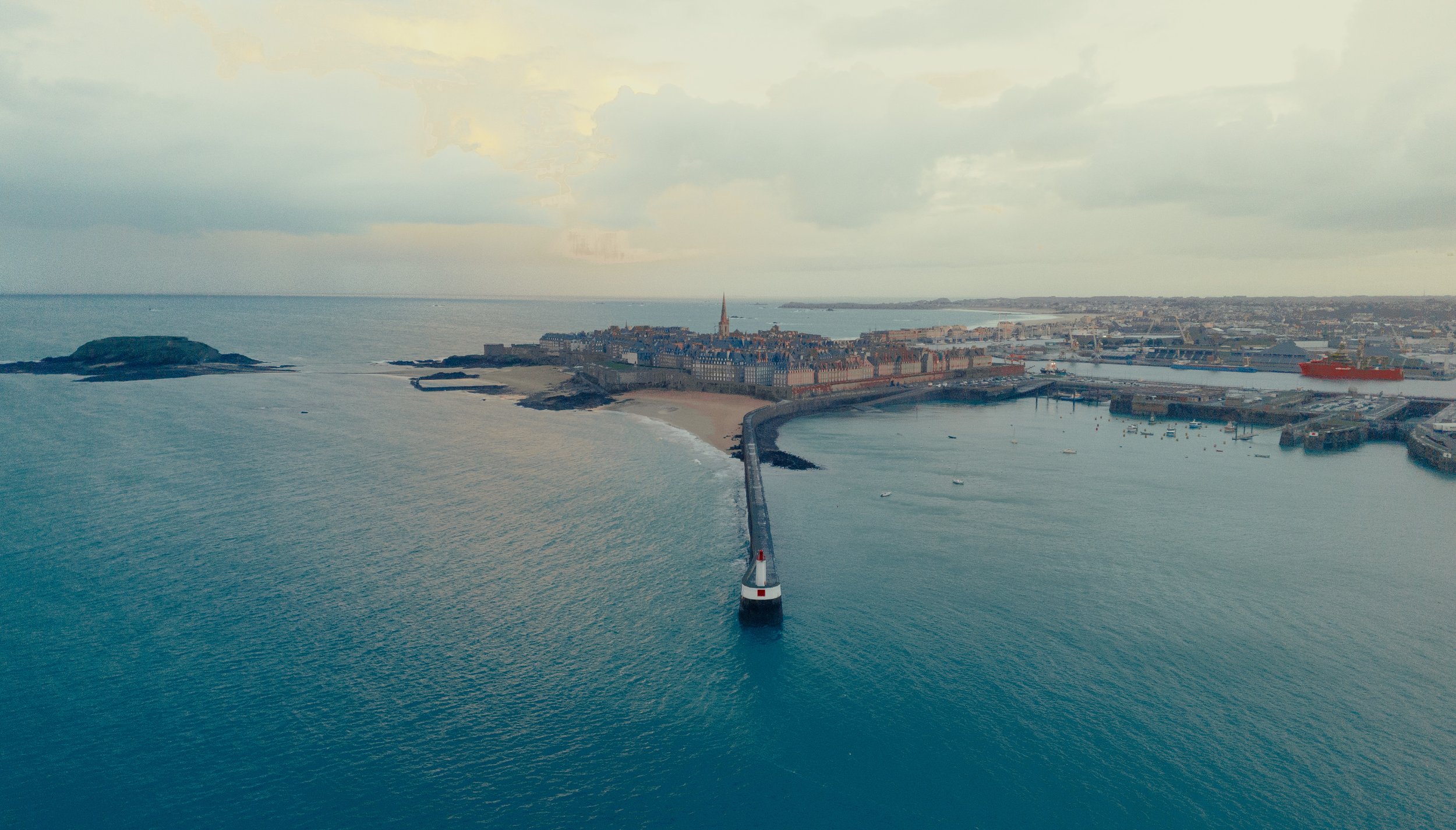 Aerial view of a coastal city with a lighthouse on a breakwater extending into the sea, a harbor with boats, historic buildings, a church with a tall spire, and a cloudy sky.