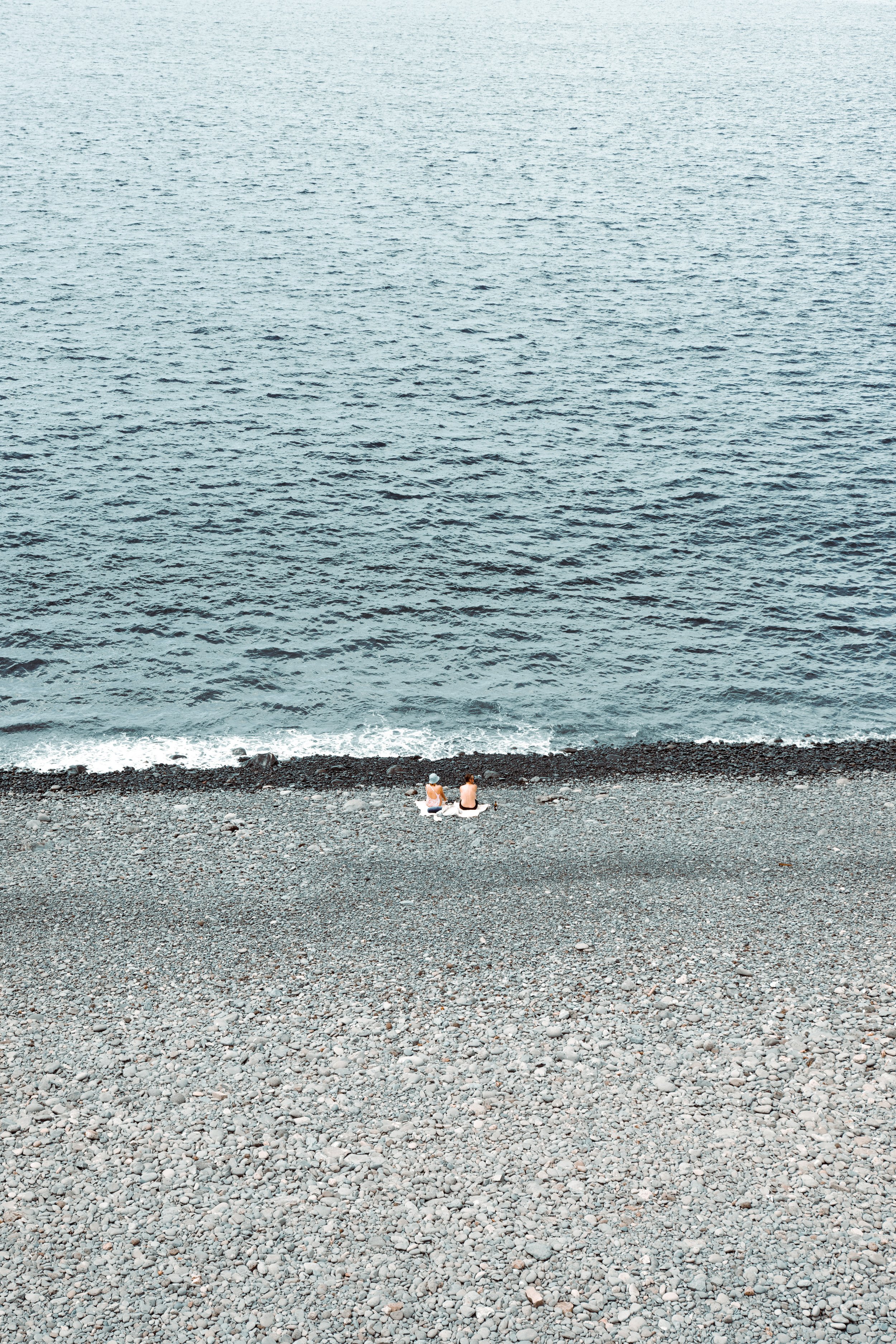 Two people sitting on a pebble beach facing the ocean.