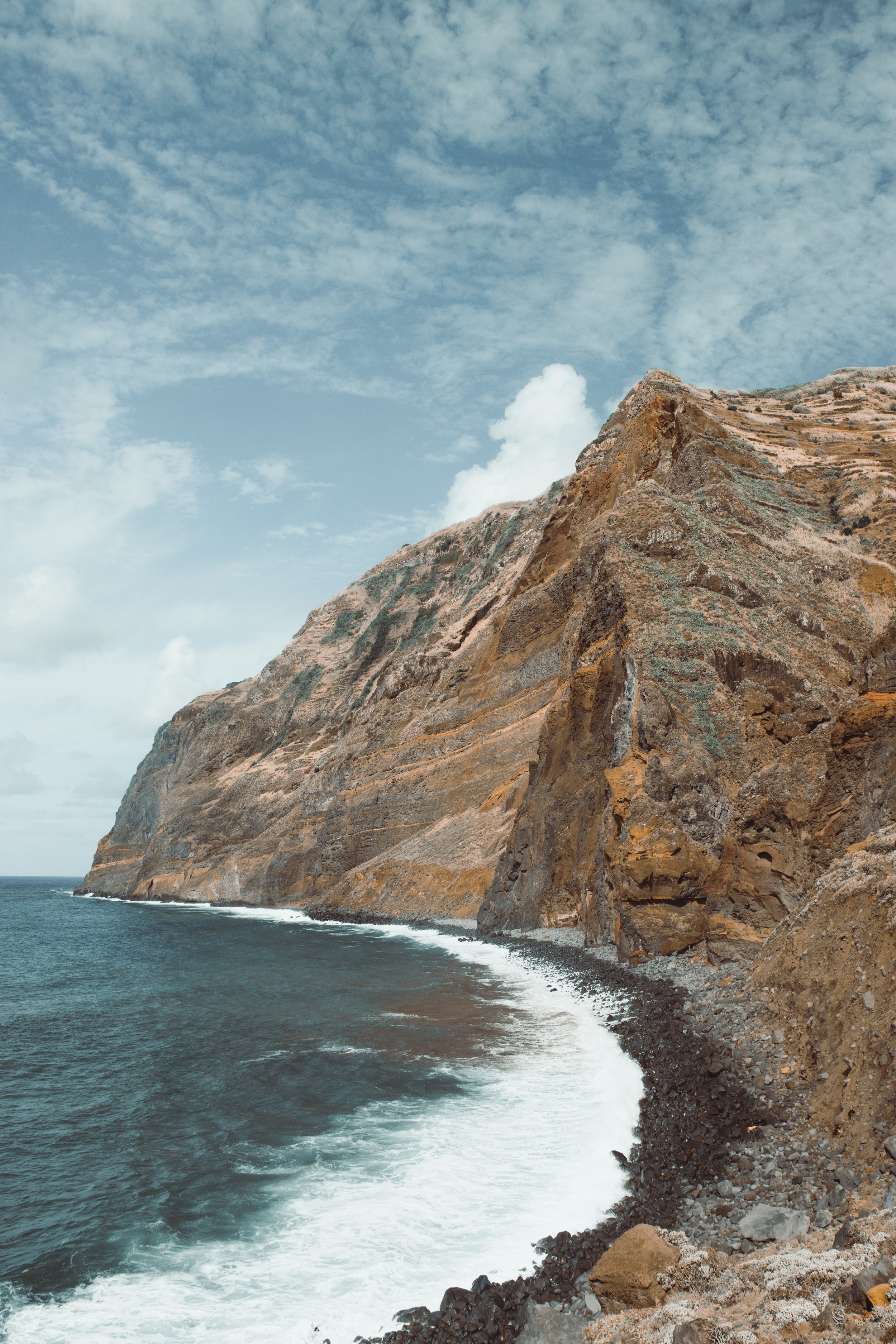 Cliffside coastline with rugged, brownish rock face and ocean waves crashing along the base under a cloudy sky.