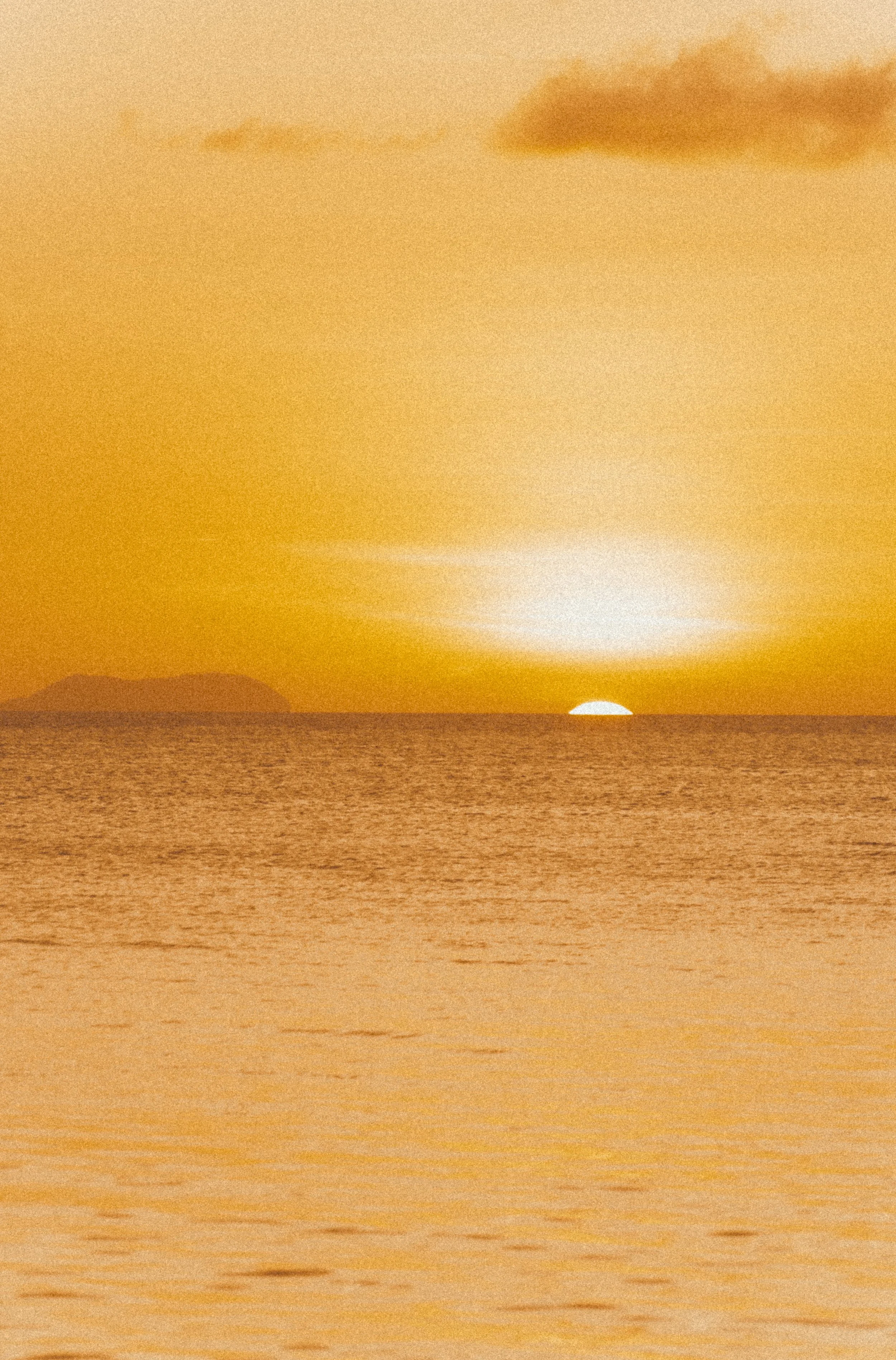 Sunset over the ocean with a partially visible island in the distance and a yellow-orange sky.