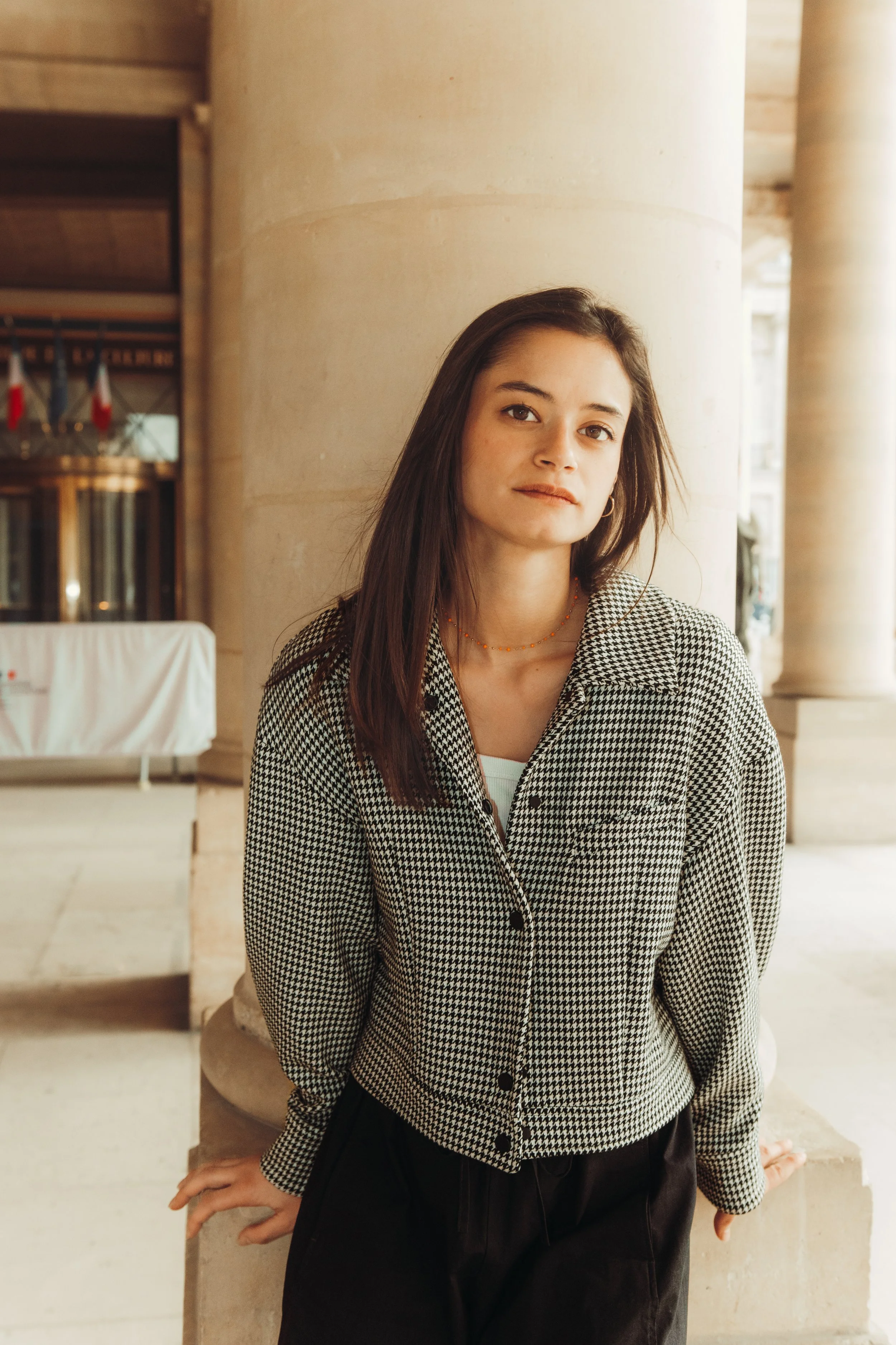 A young woman with straight brown hair wearing a houndstooth checkered jacket and black pants, standing outdoors near large stone columns.