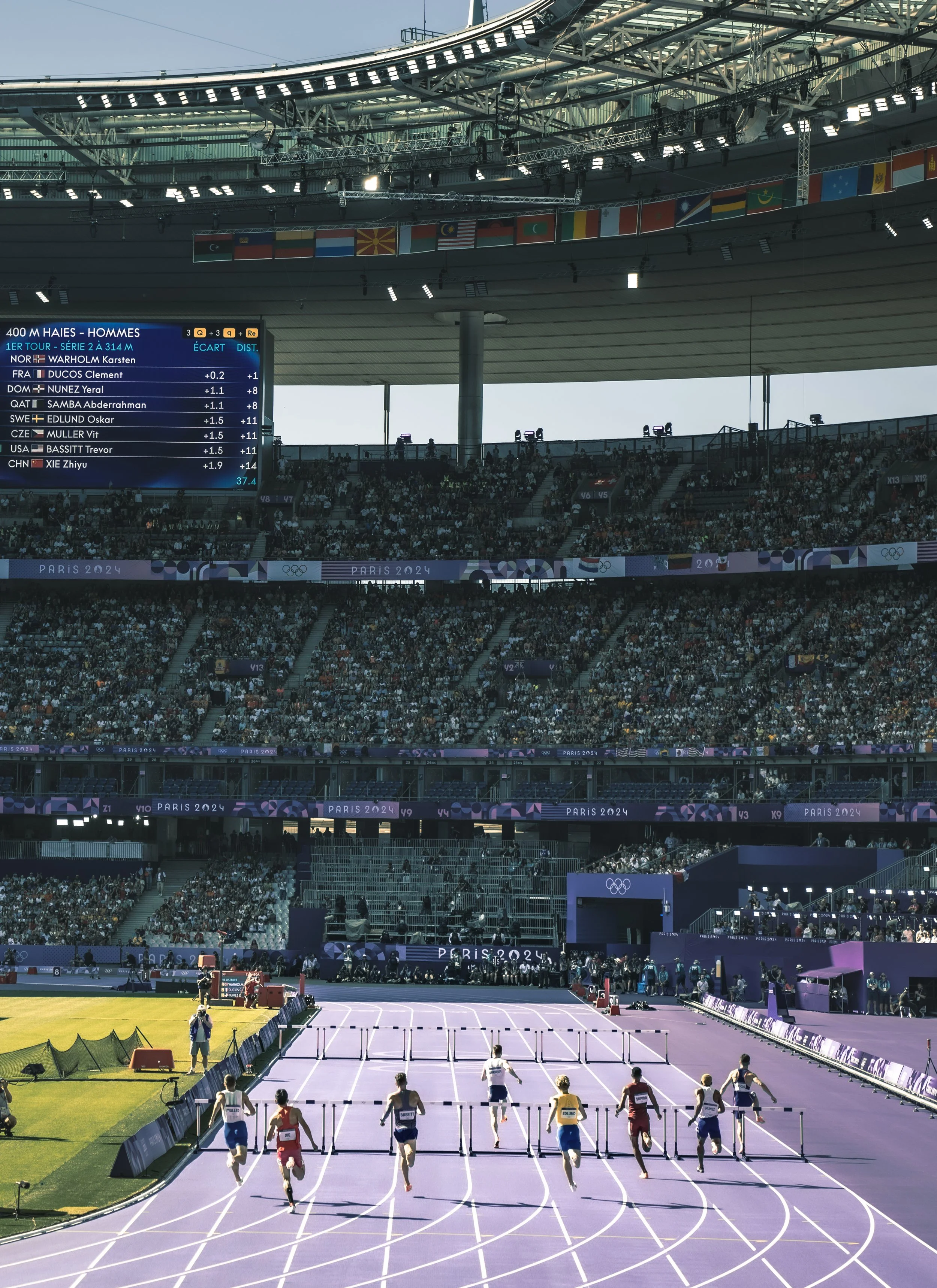 An indoor track at the Paris 2024 Olympics with athletes running in a race. A large crowd is in the stands, and flags from various countries are hanging from the ceiling. The scoreboard shows the race details and competitors.