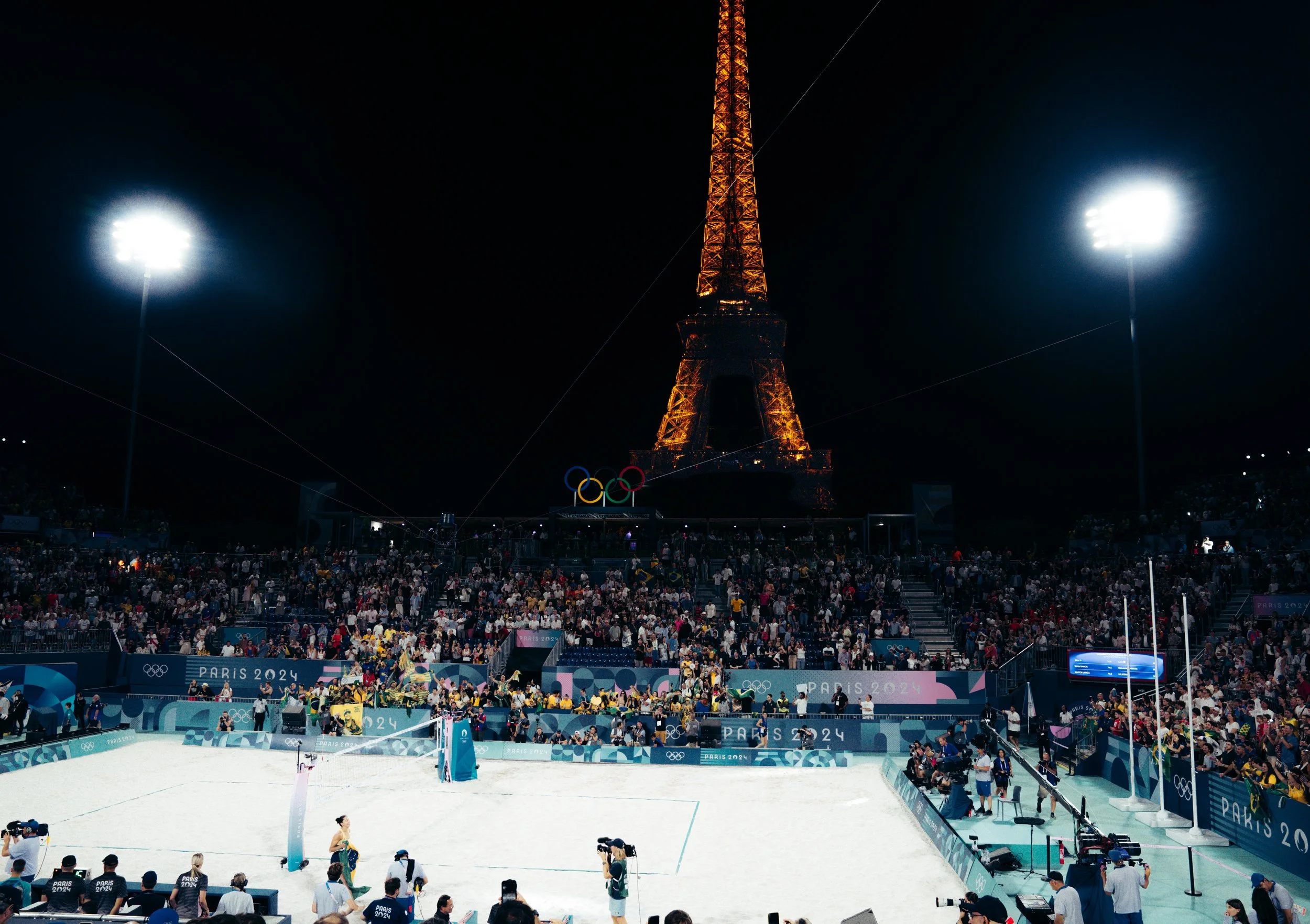 Nighttime view of a beach volleyball game at the Paris 2024 Olympics, with the Eiffel Tower illuminated in the background and large crowd spectators.