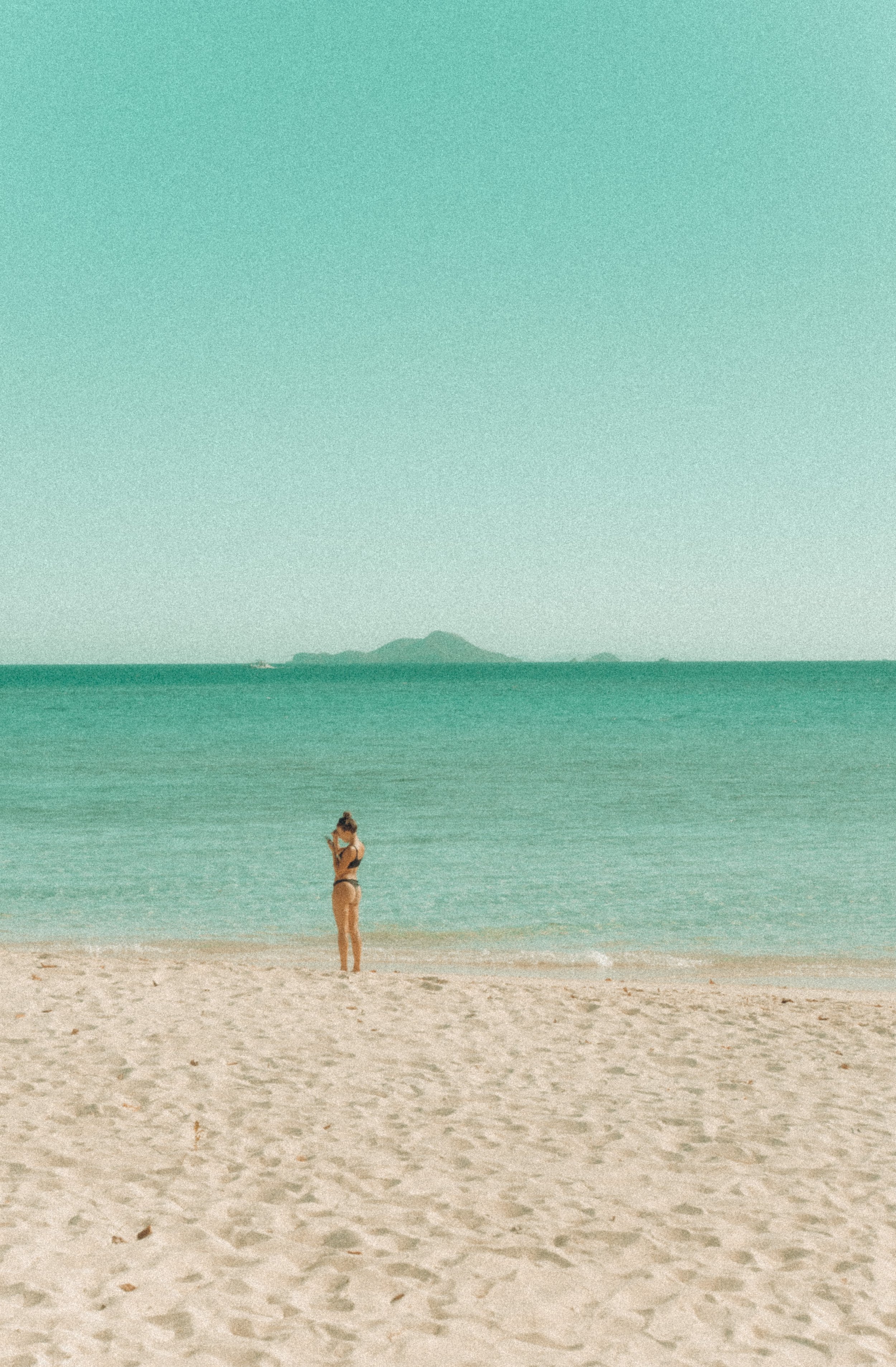 A woman in a bikini standing on a sandy beach, looking at her phone with the ocean and an island in the background.