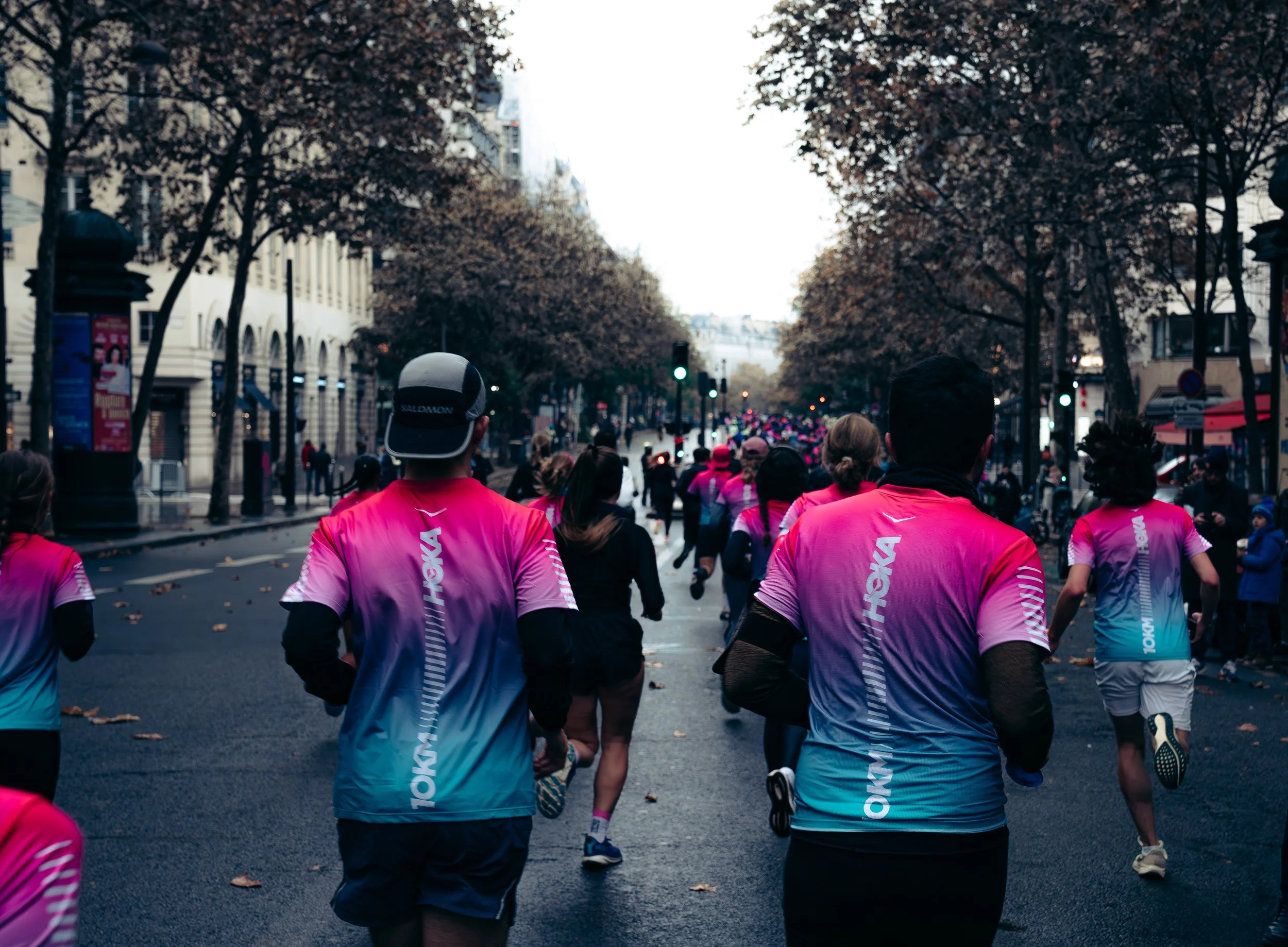 People running in a marathon on a city street lined with trees and buildings, wearing pink and blue athletic shirts.