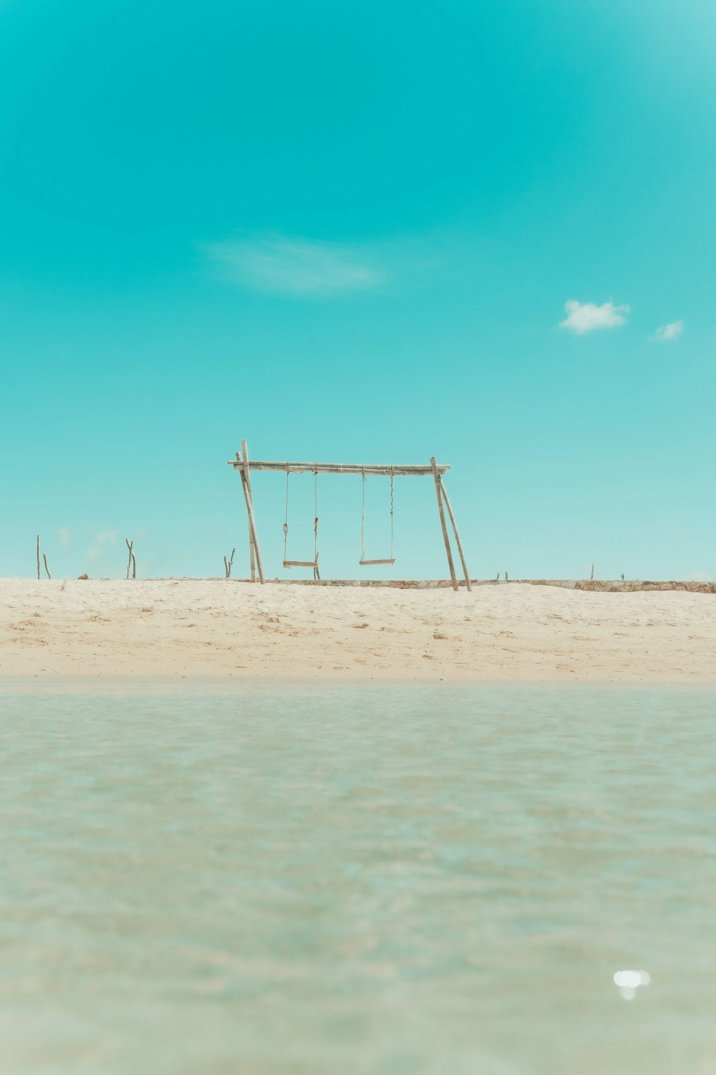 Empty wooden swingset on sandy beach with calm water in foreground and clear blue sky.