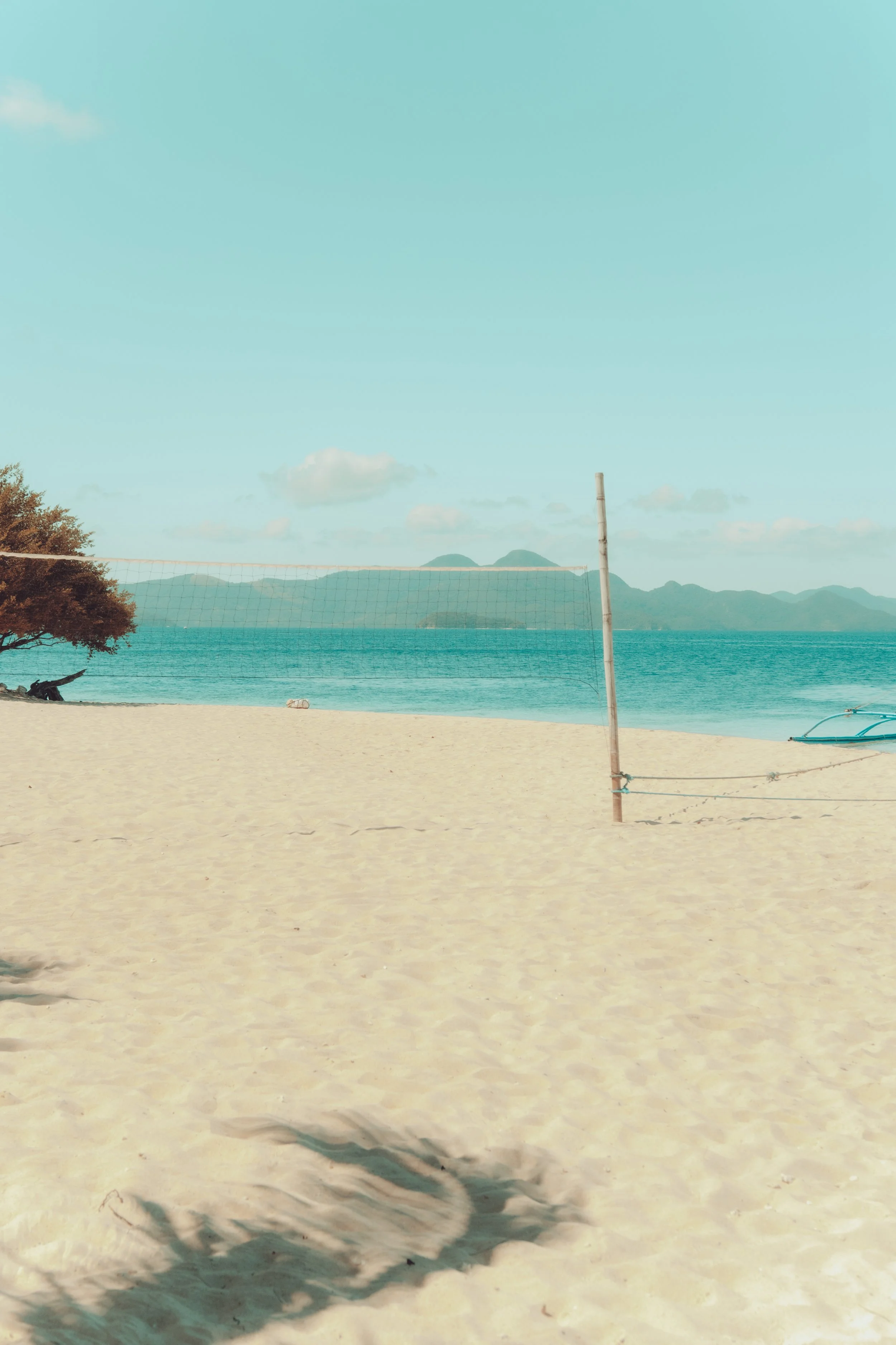 A sandy beach with a volleyball net set up, a tree casting shadows, calm ocean waters, and distant mountains under a partly cloudy sky.