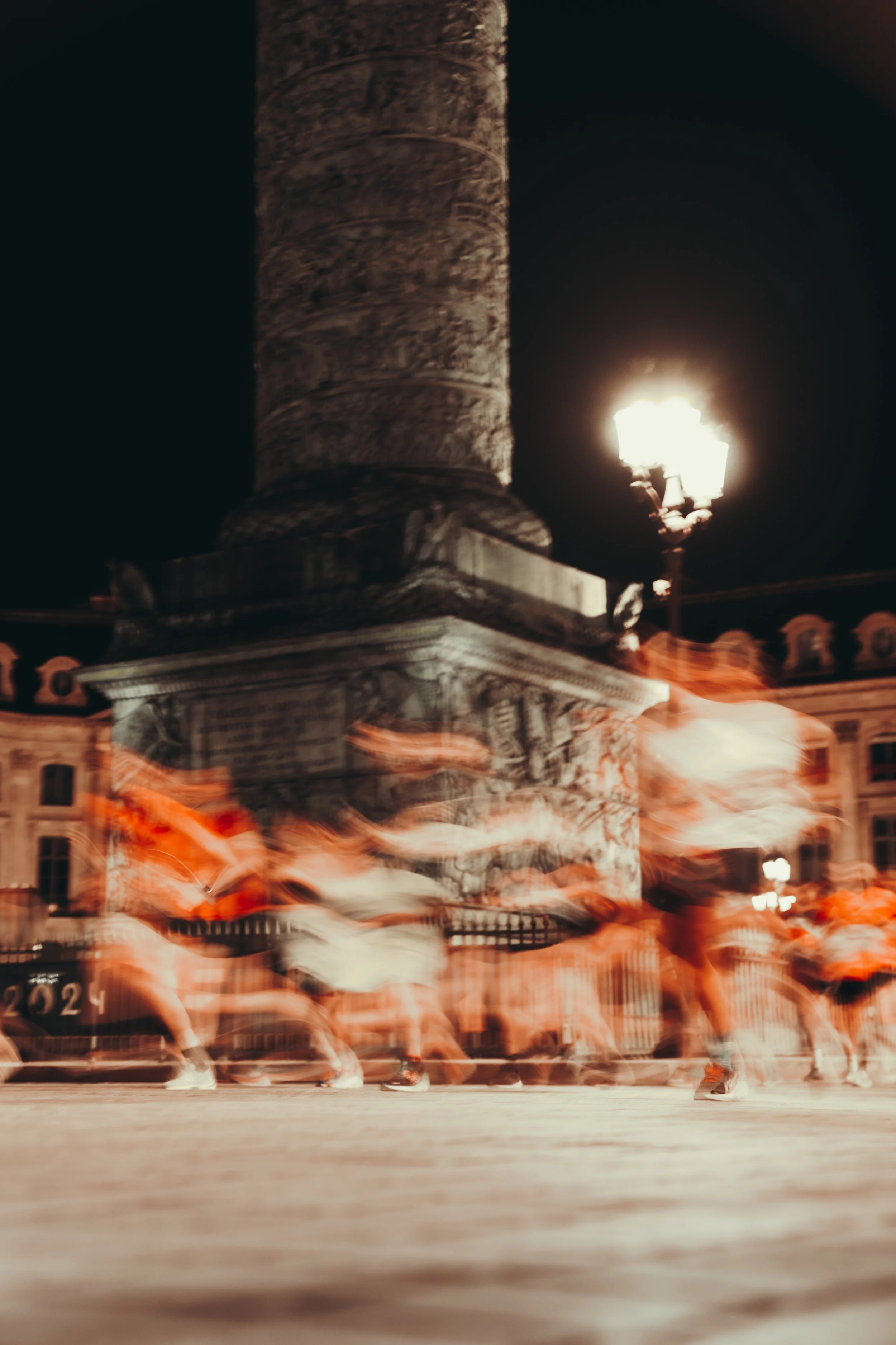Nighttime street scene with blurred motion of people walking past a historic stone monument and a lamppost illuminating the area.