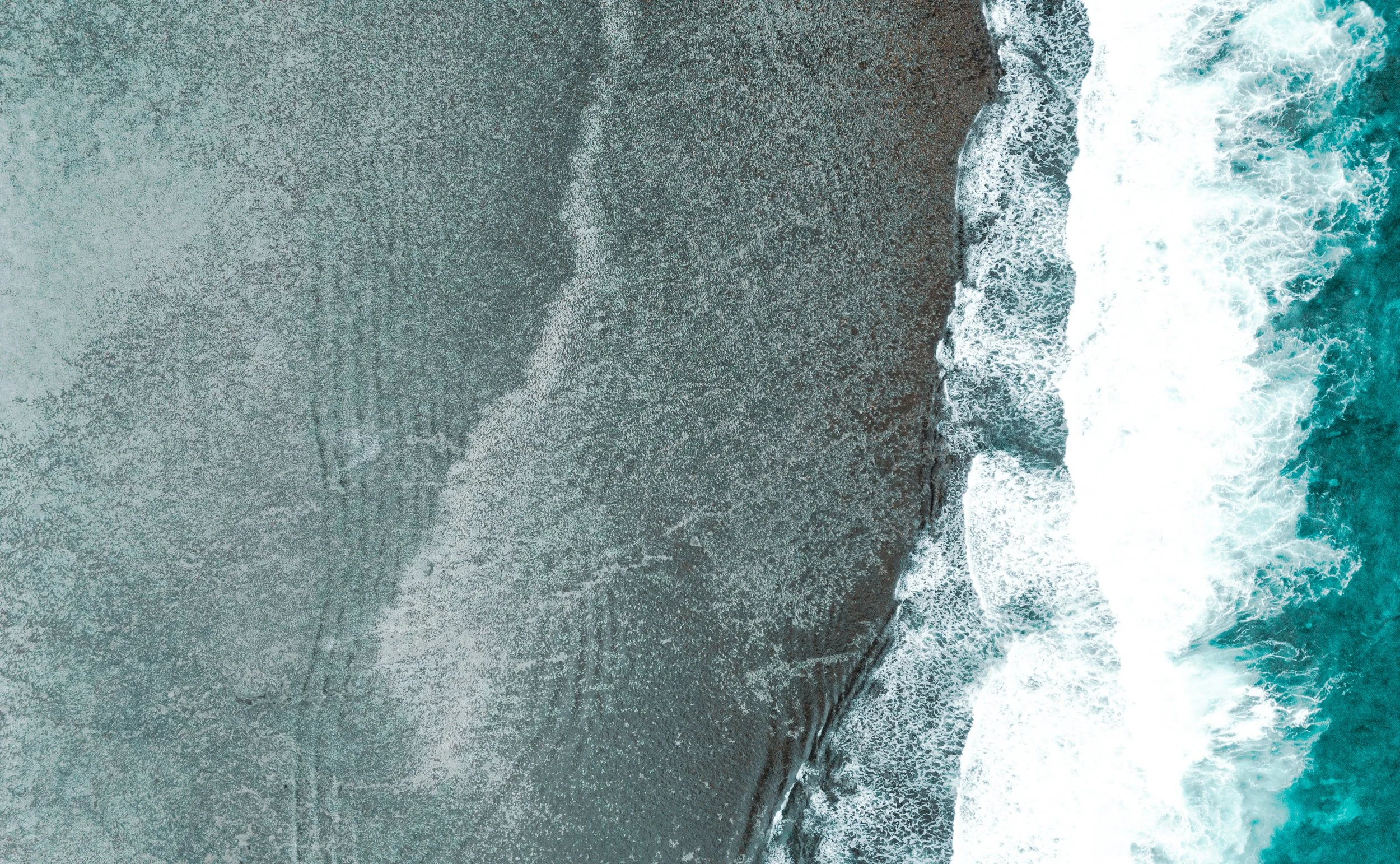 An aerial view of a dark sandy beach with gentle ocean waves crashing onto the shore.