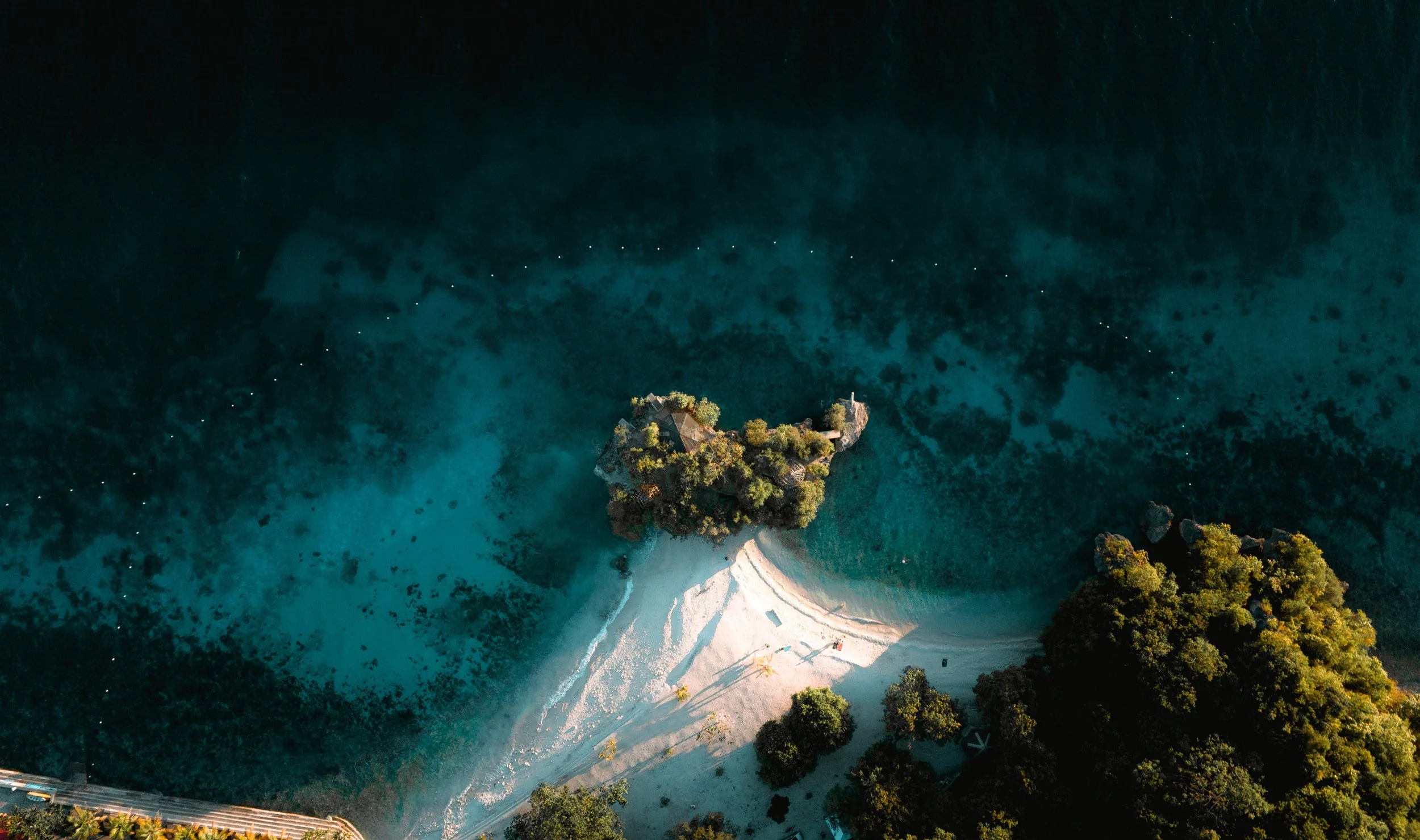 Aerial view of a small island with lush greenery, sandy beach, and a rocky outcrop in the ocean, surrounded by clear blue water.