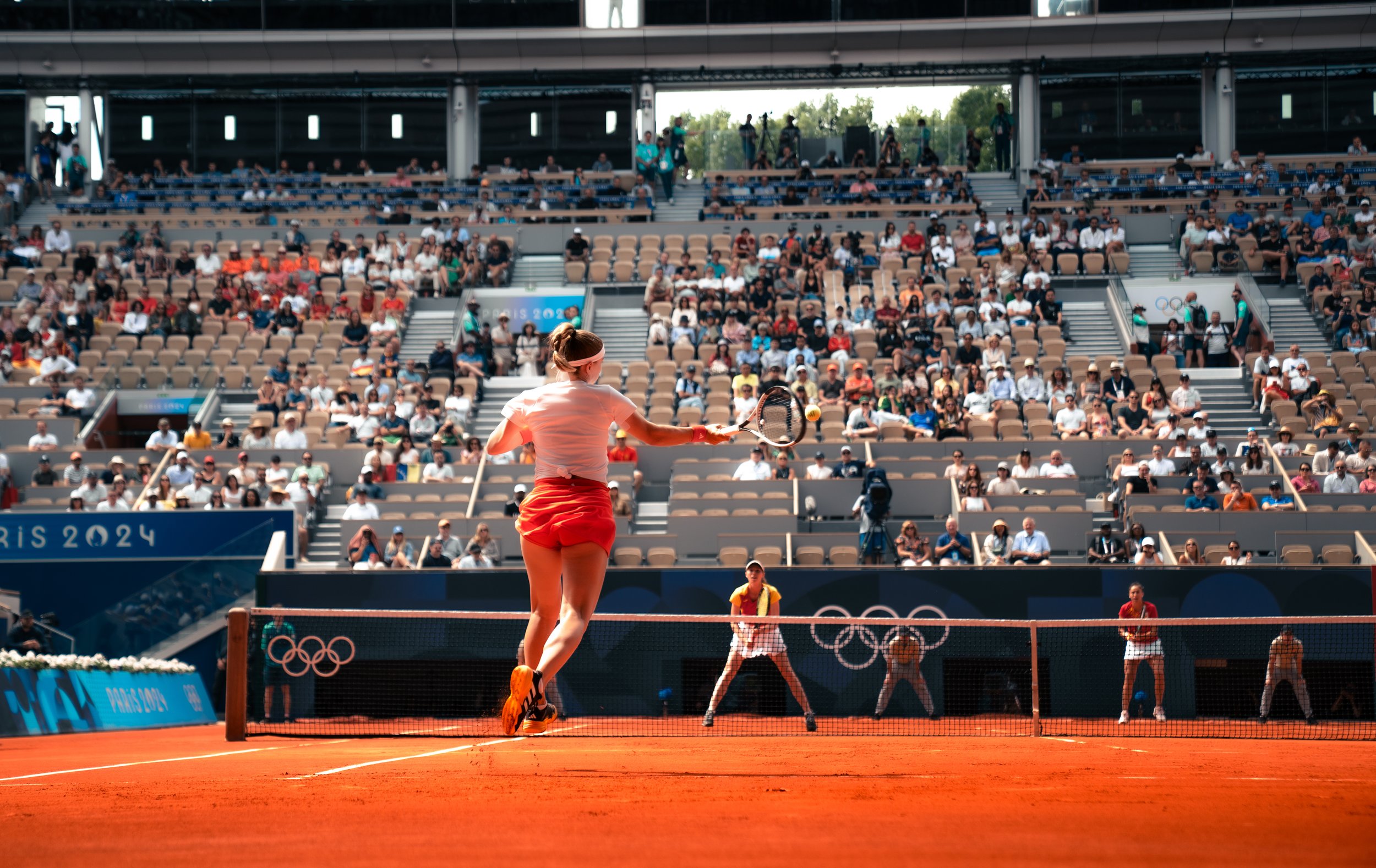A female tennis player in an orange skirt and white top hits a tennis ball on a clay court during the Olympics, with spectators in the stadium watching.