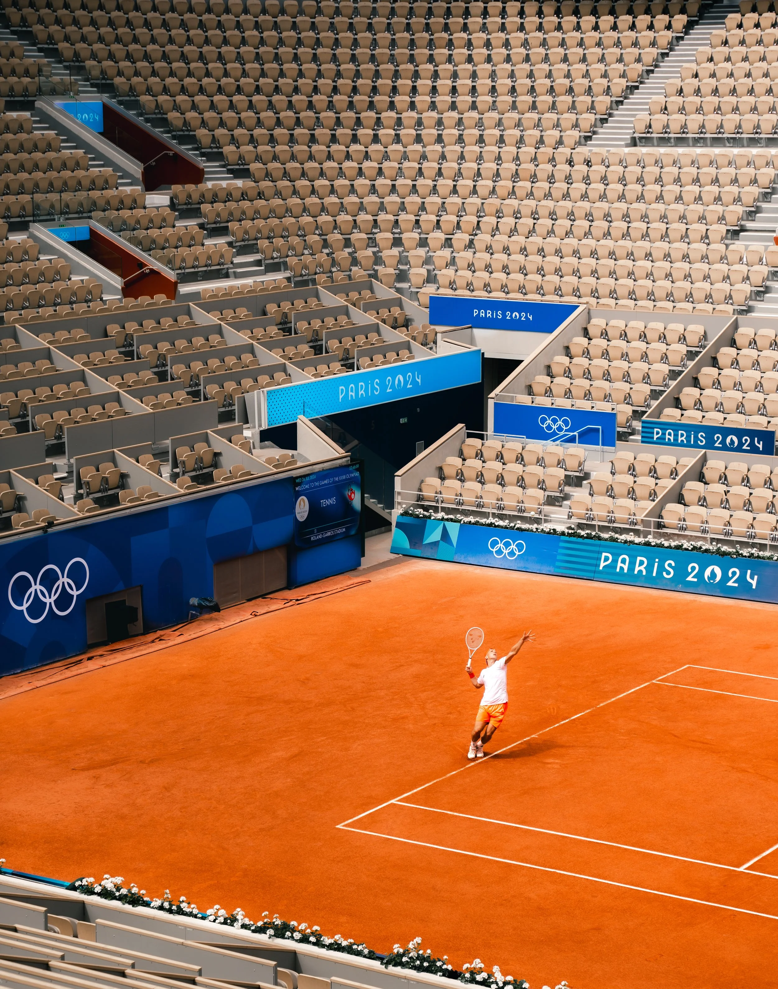 A solitary tennis player on an orange clay court at the Paris 2024 Olympics, with empty stadium seating surrounding the court.