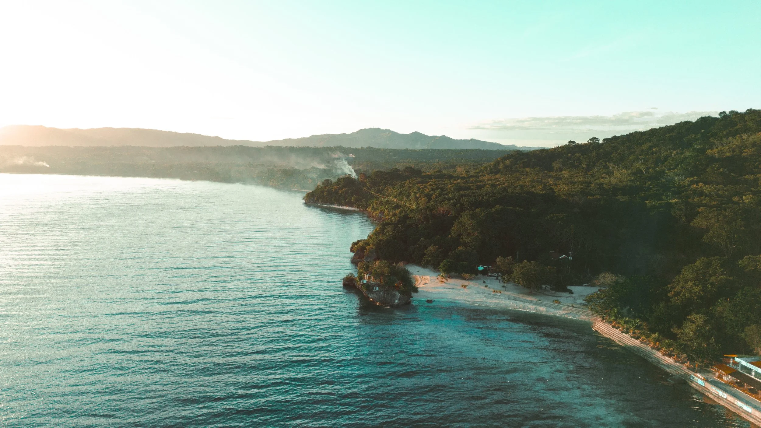 Aerial view of a coastal landscape with a beach, dense forest, and ocean water, with hills in the background.