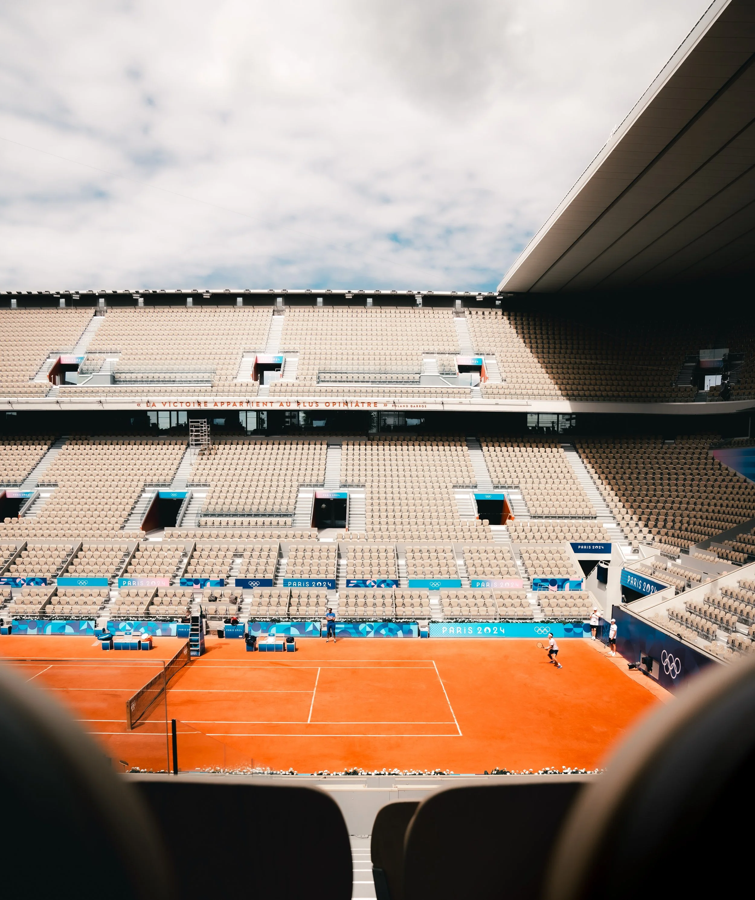 Empty tennis court at the Paris 2024 Olympics with stadium seats and sky overhead.