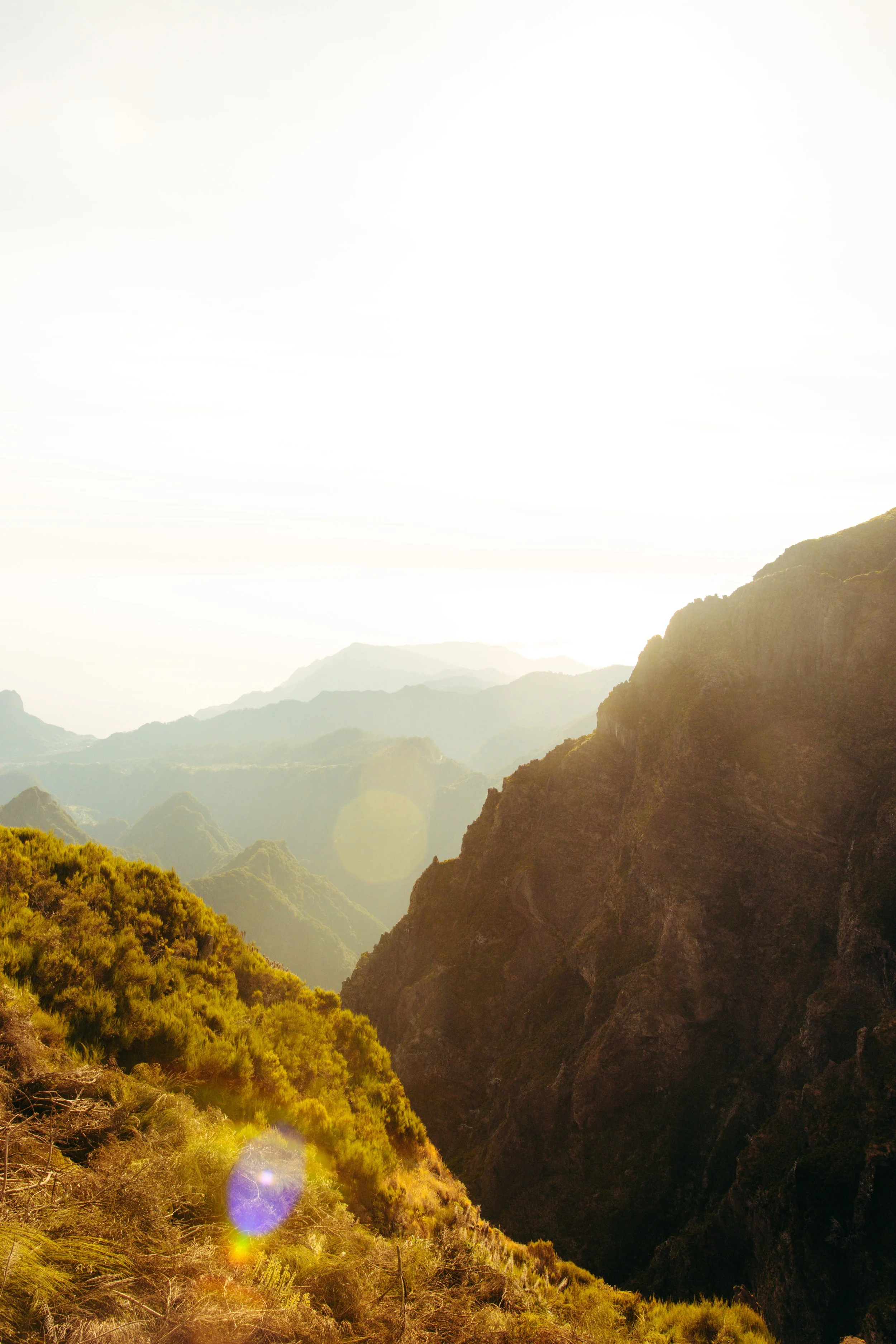 Sunlit mountain landscape with rugged cliffs and lush green vegetation in the foreground, hazy mountain ranges in the background, and a bright sky overhead.
