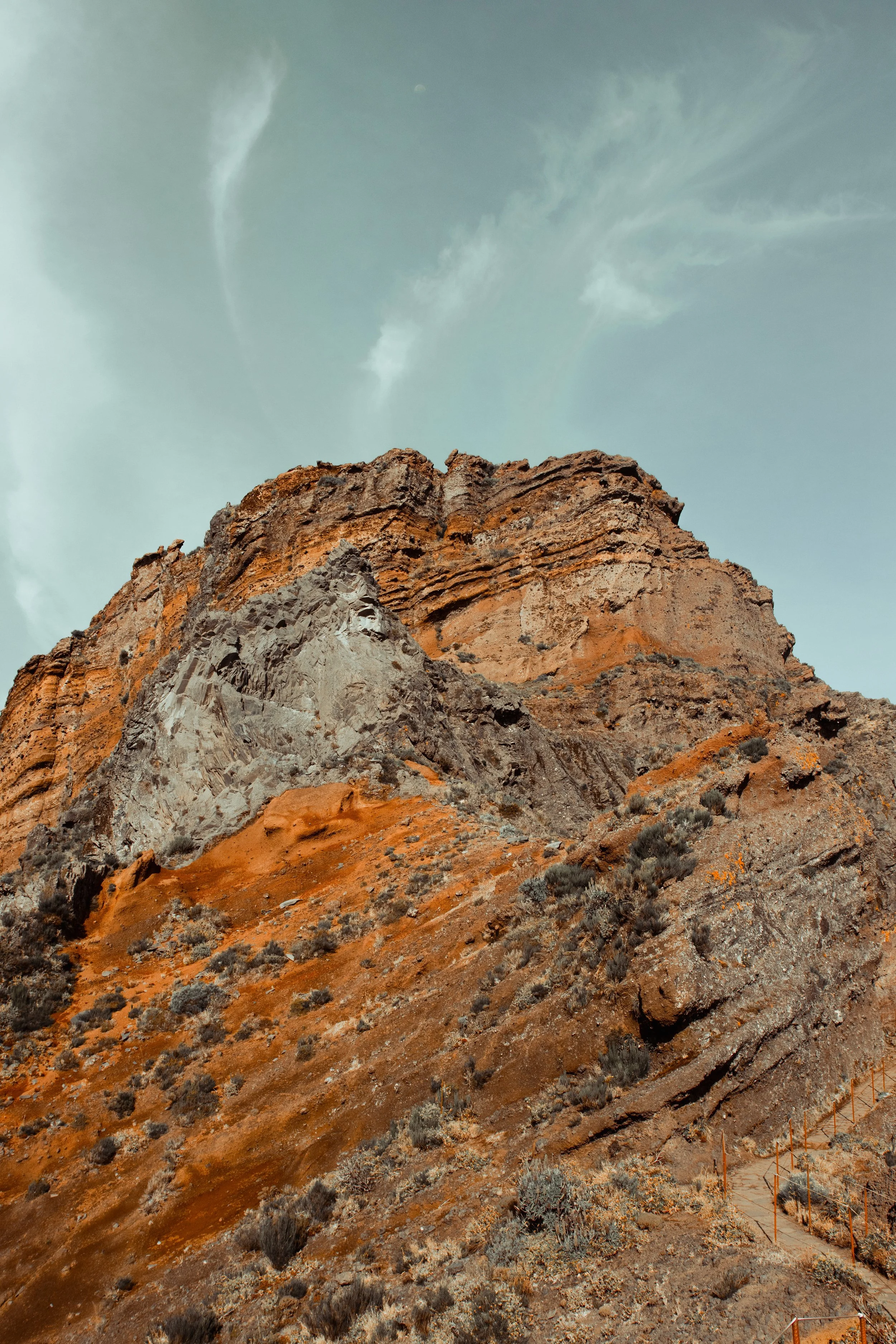 A rugged mountain with orange and brown rocky terrain and sparse vegetation, with a partly cloudy sky overhead.