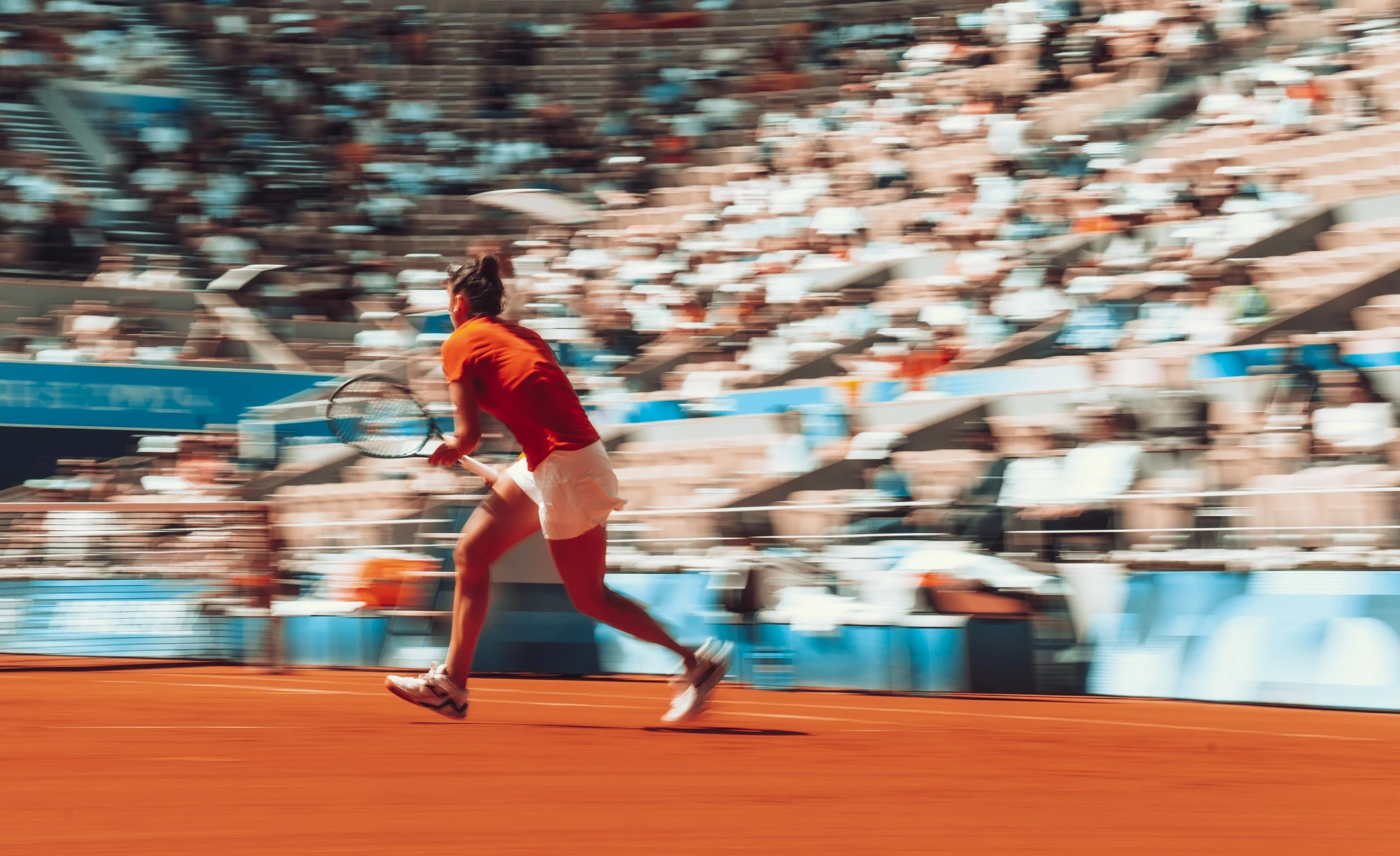 A female tennis player running on a clay court during a match as the audience watches from the stands.