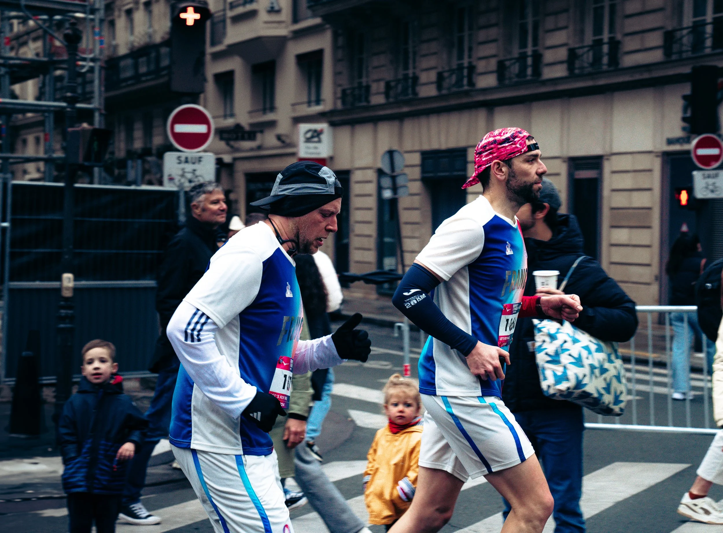 Two male runners participating in a marathon, wearing blue and white sportswear, running on a city street with pedestrians and spectators around.