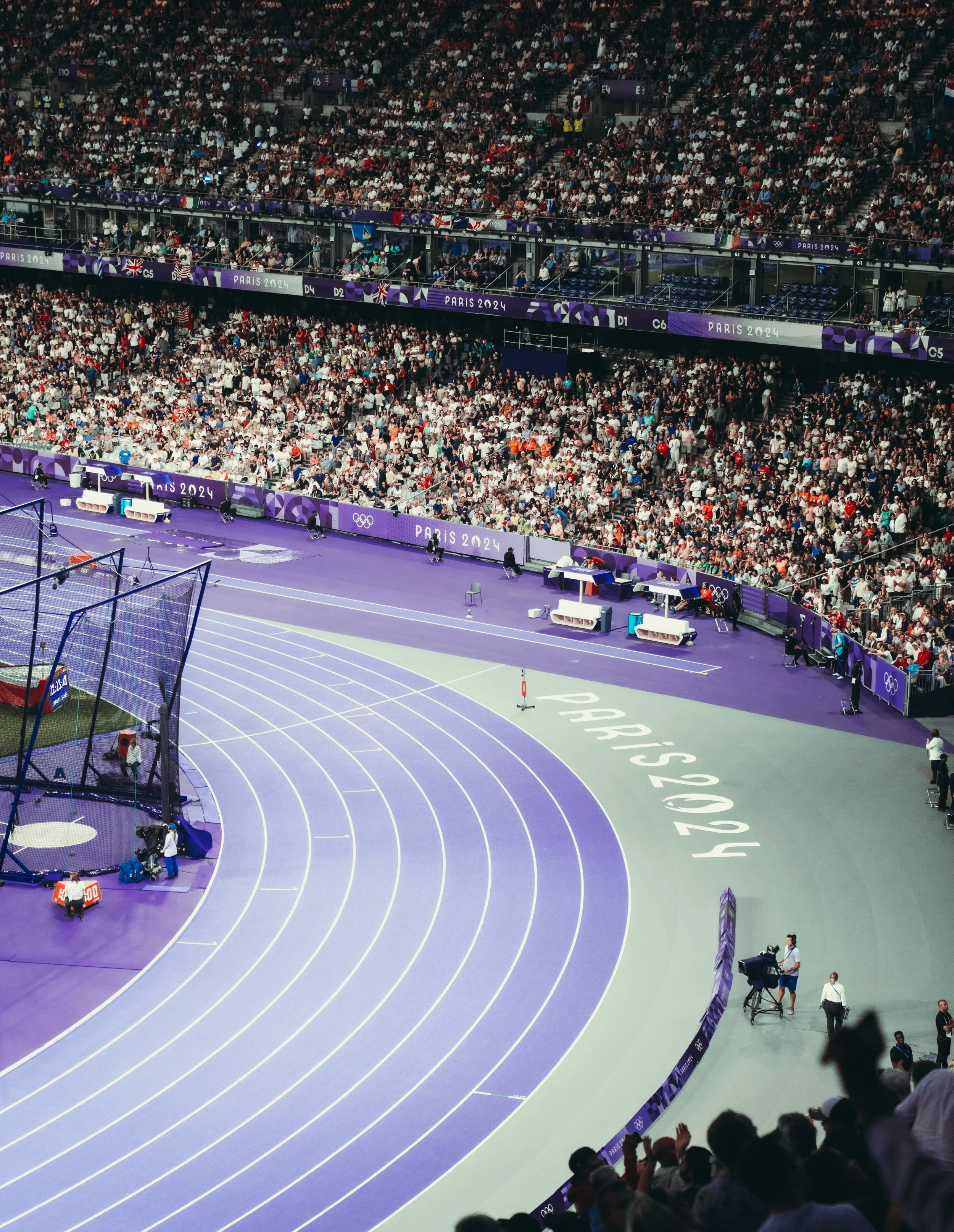 Indoor Olympic track and field stadium filled with spectators, with a purple running track and around 4 lanes visible. There are various officials and crew around, and the stadium has banners reading 'Paris 2024' and the Olympic rings.