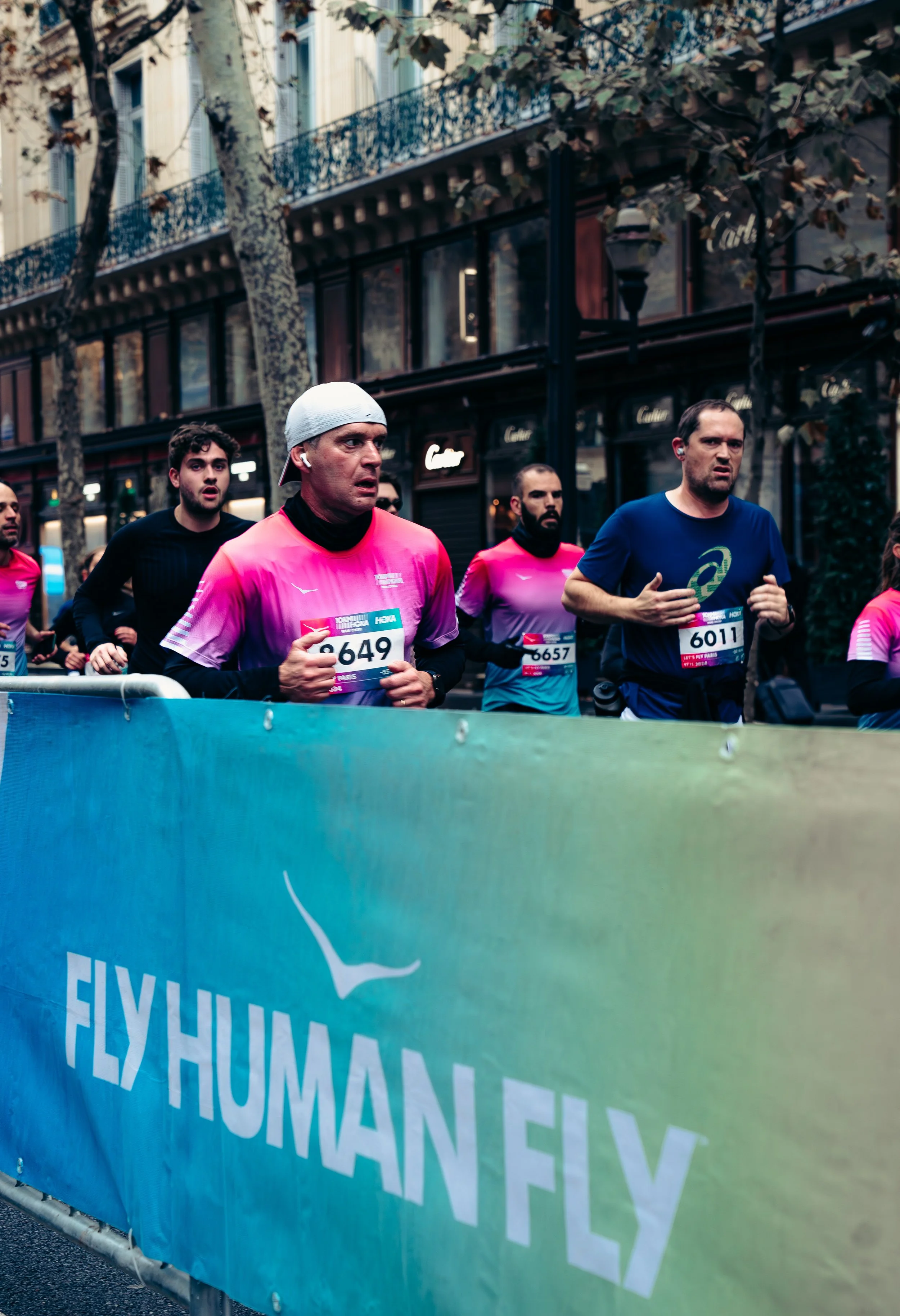 Group of marathon runners running past a blue banner that reads 'FLYHUMAN FLY' during a race in an urban setting, with trees and buildings in the background.